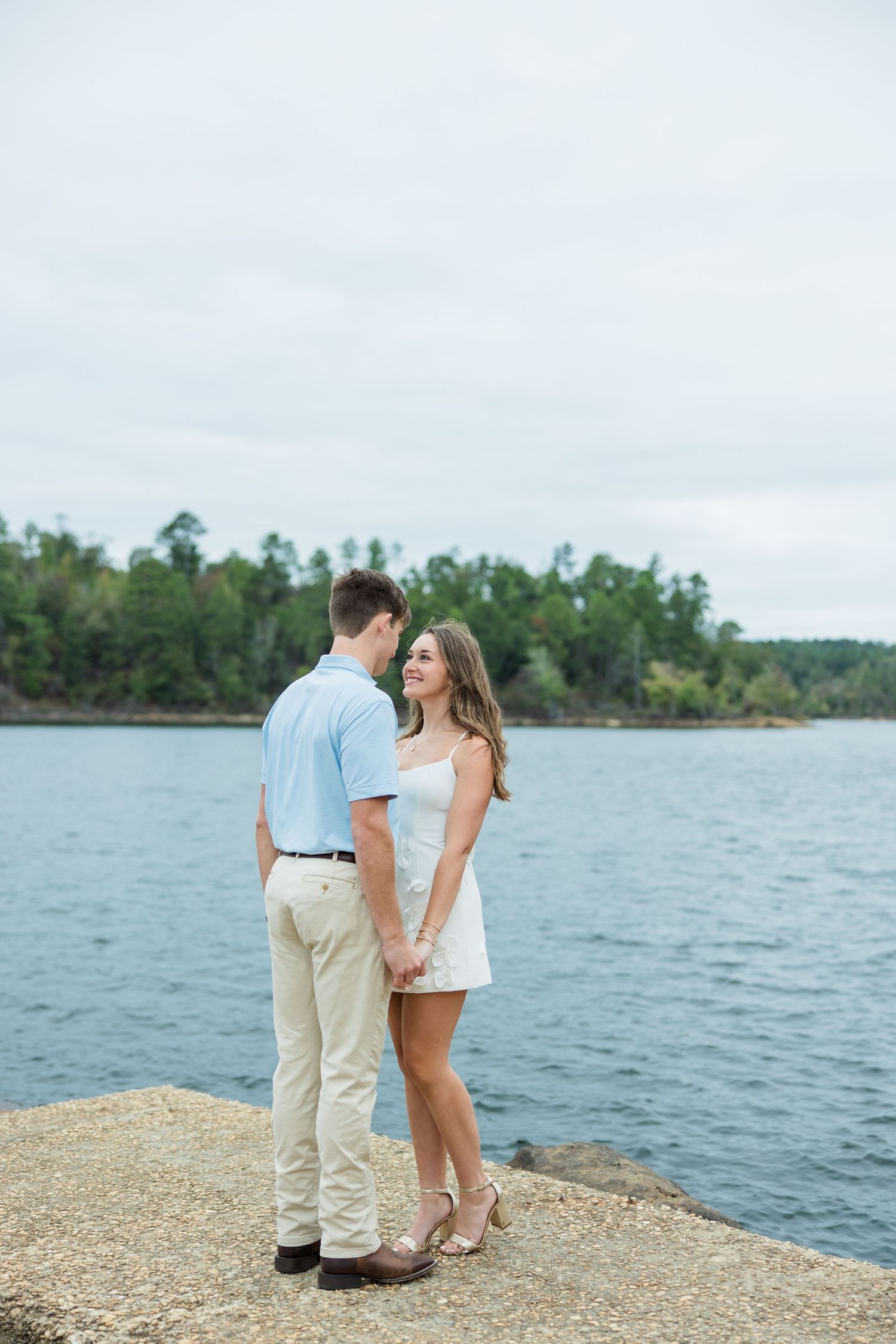 Smiling couple holding hands and facing each other on a stone lakeside ledge with calm water and green trees behind them.