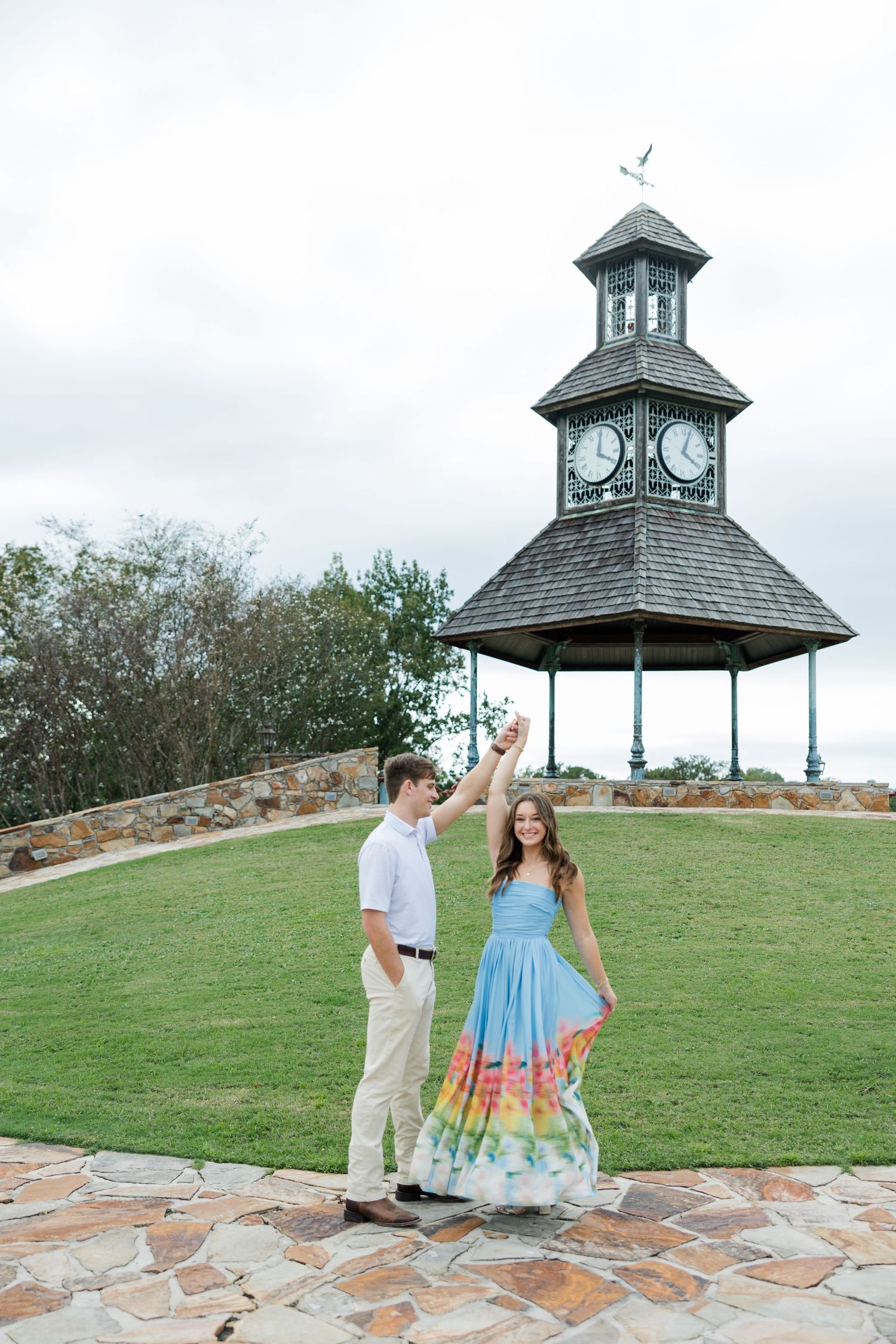 Couple dancing on a stone path in front of a grassy hill and a clock tower gazebo during an engagement photo session.