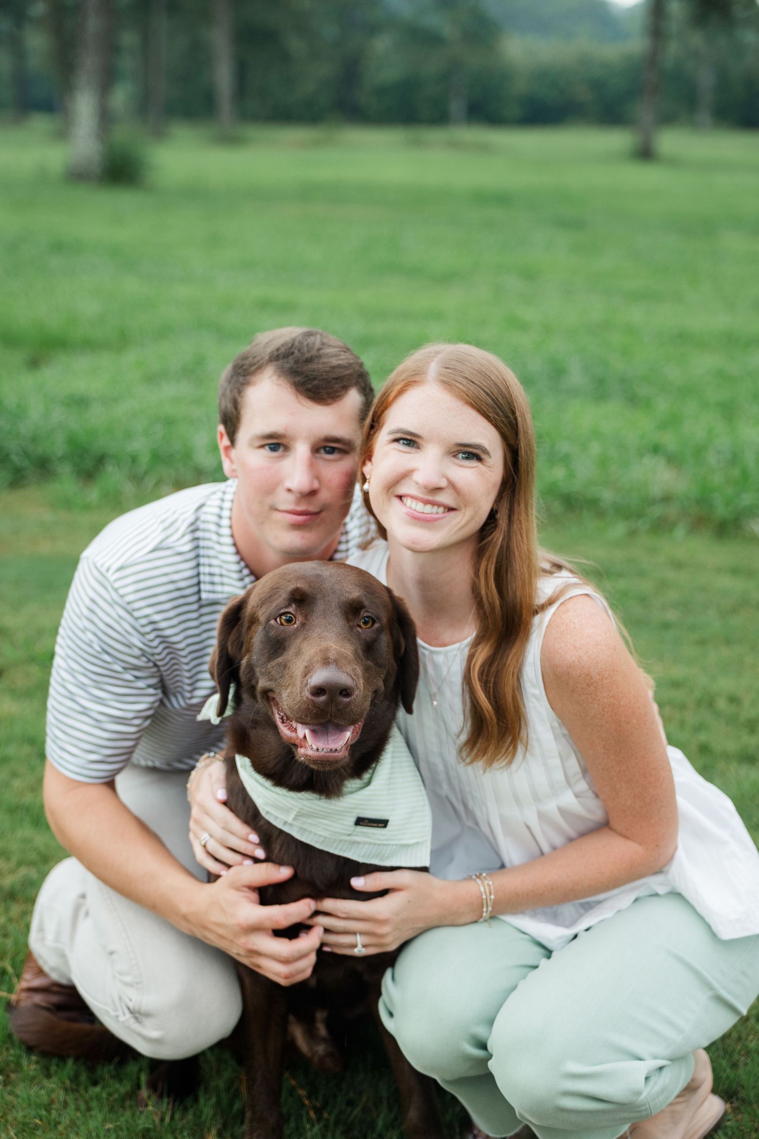 Smiling couple crouching with their chocolate Labrador dog on a green grass field.