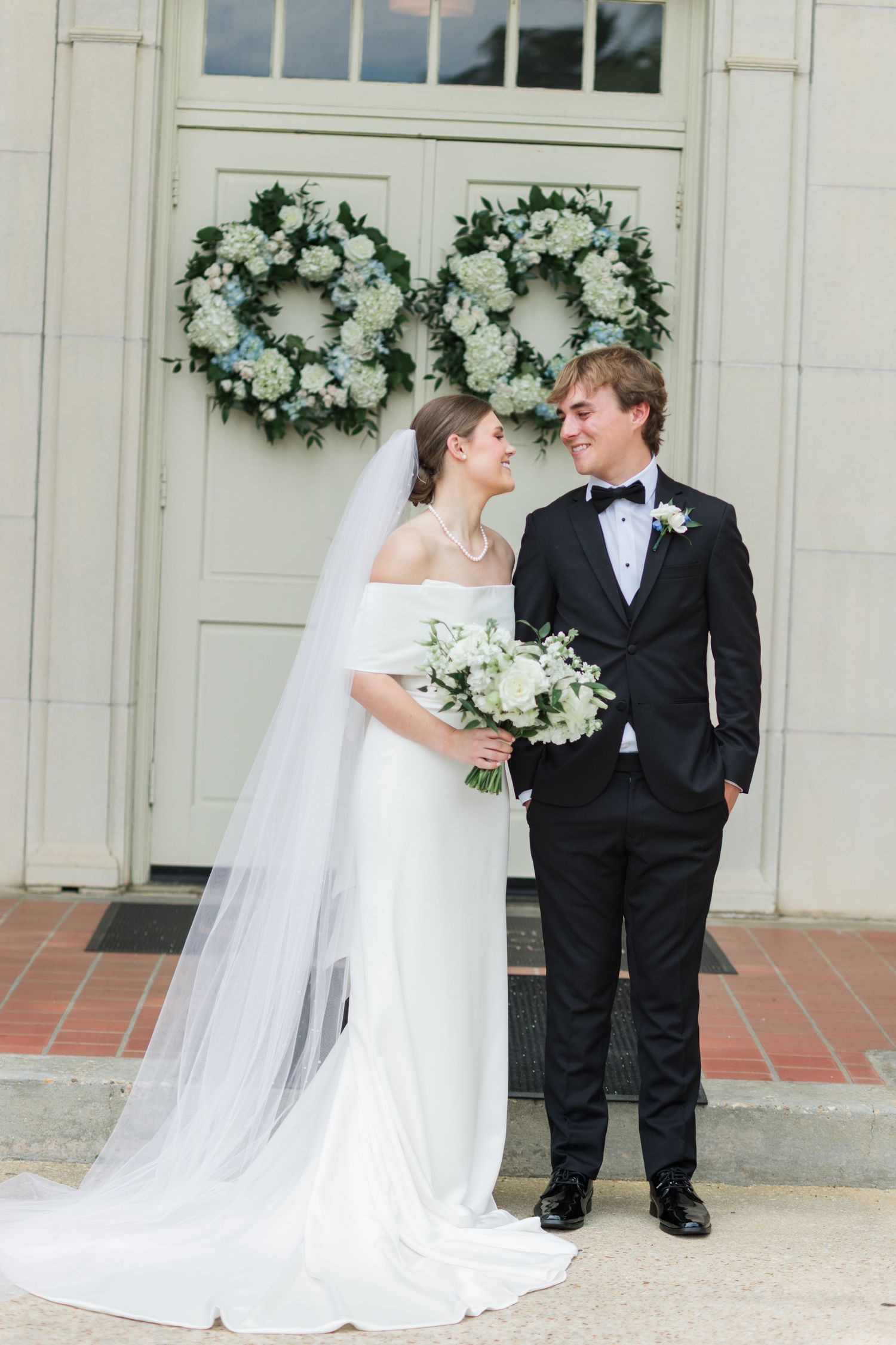 Bride and groom pose before church doors adorned with white and blue hydrangea wreaths.