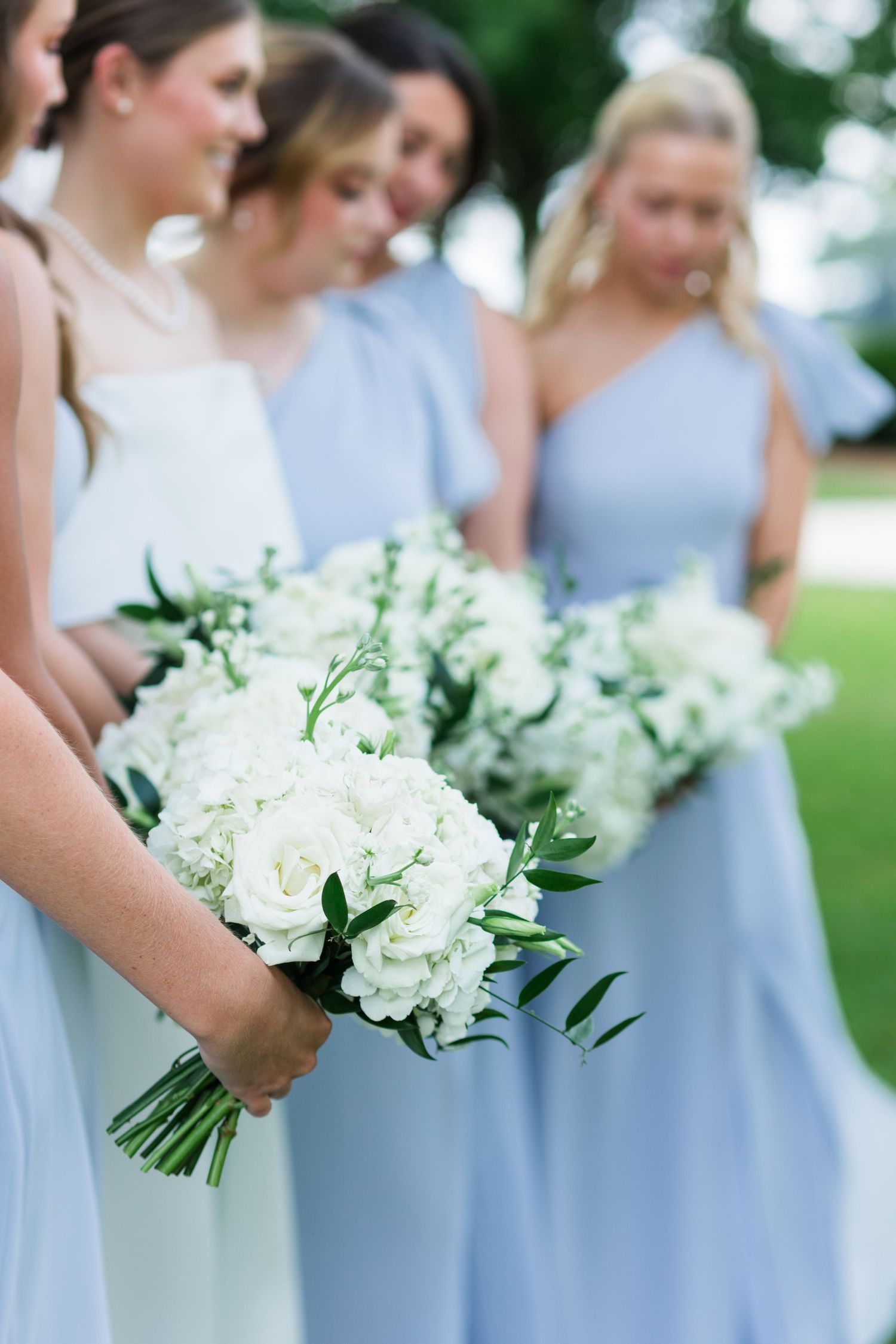 Bridesmaids in light blue dresses holding white floral bouquets with greenery at wedding.