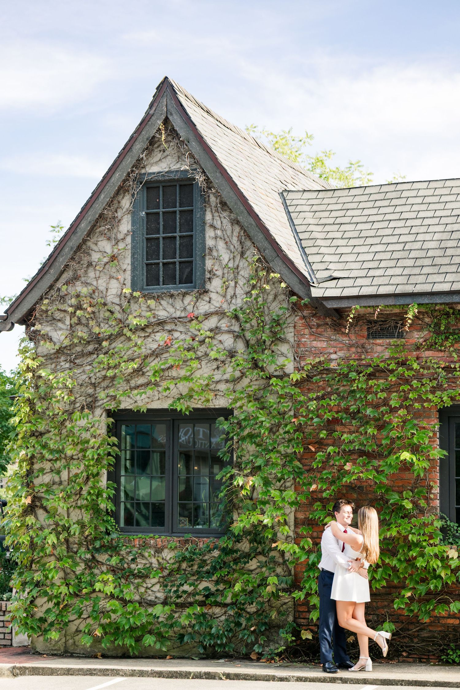 A couple shares a romantic embrace outside an ivy-covered stone and brick cottage under a blue sky.