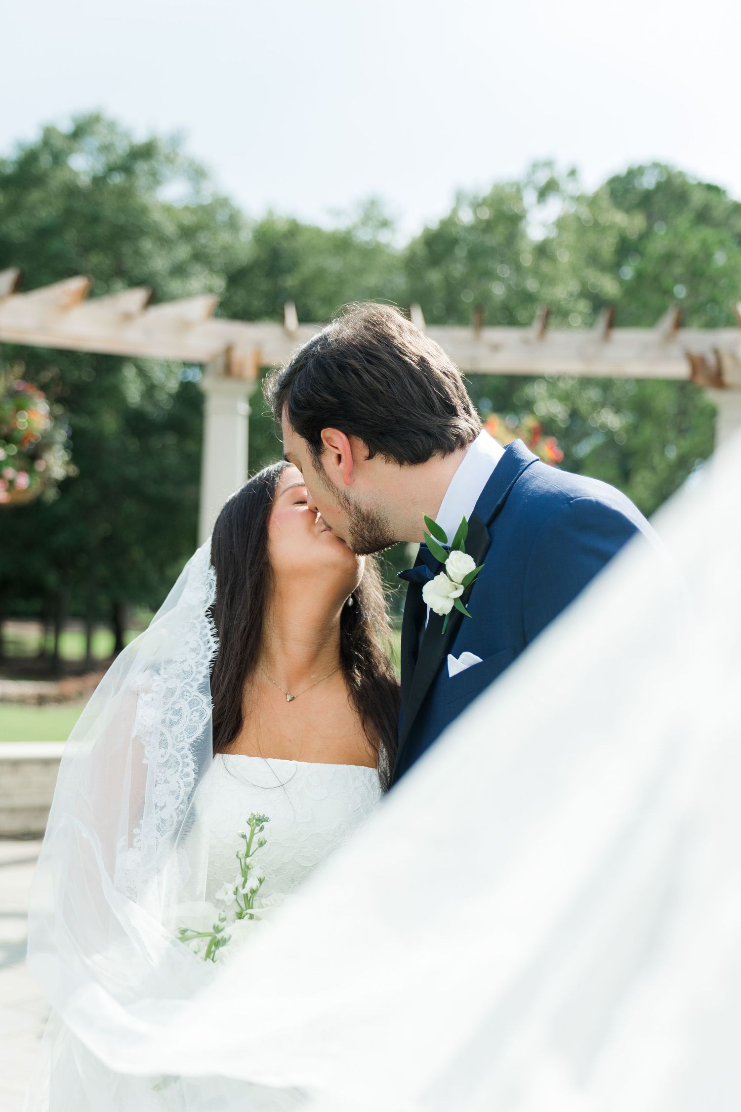 Bride and groom sharing a romantic kiss with flowing veil billowing in the breeze under a pergola.