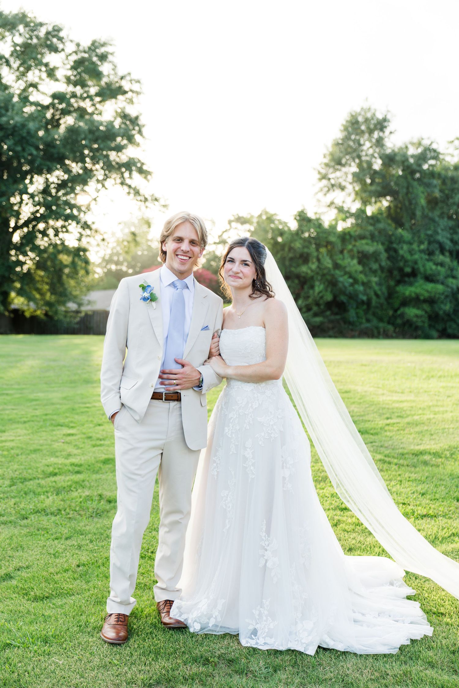 Bride and groom posing together on a green lawn at golden hour, both smiling in elegant wedding attire.