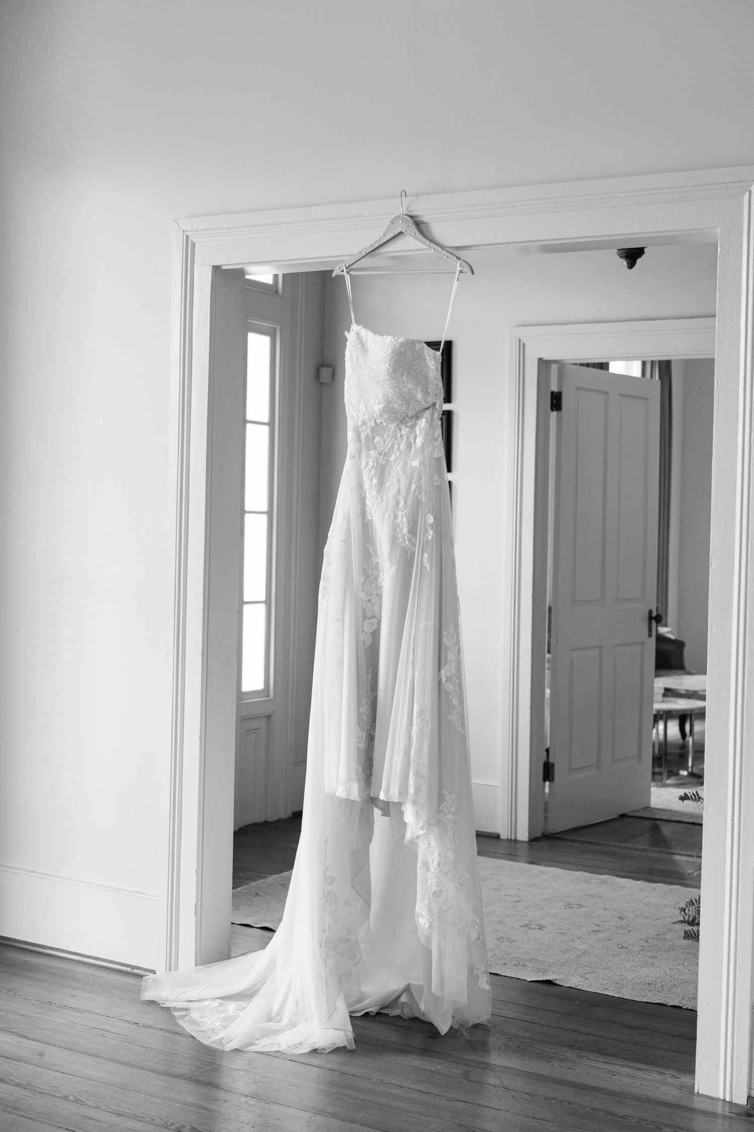 A elegant lace wedding dress hanging on a hanger in a bright white room with hardwood floors.