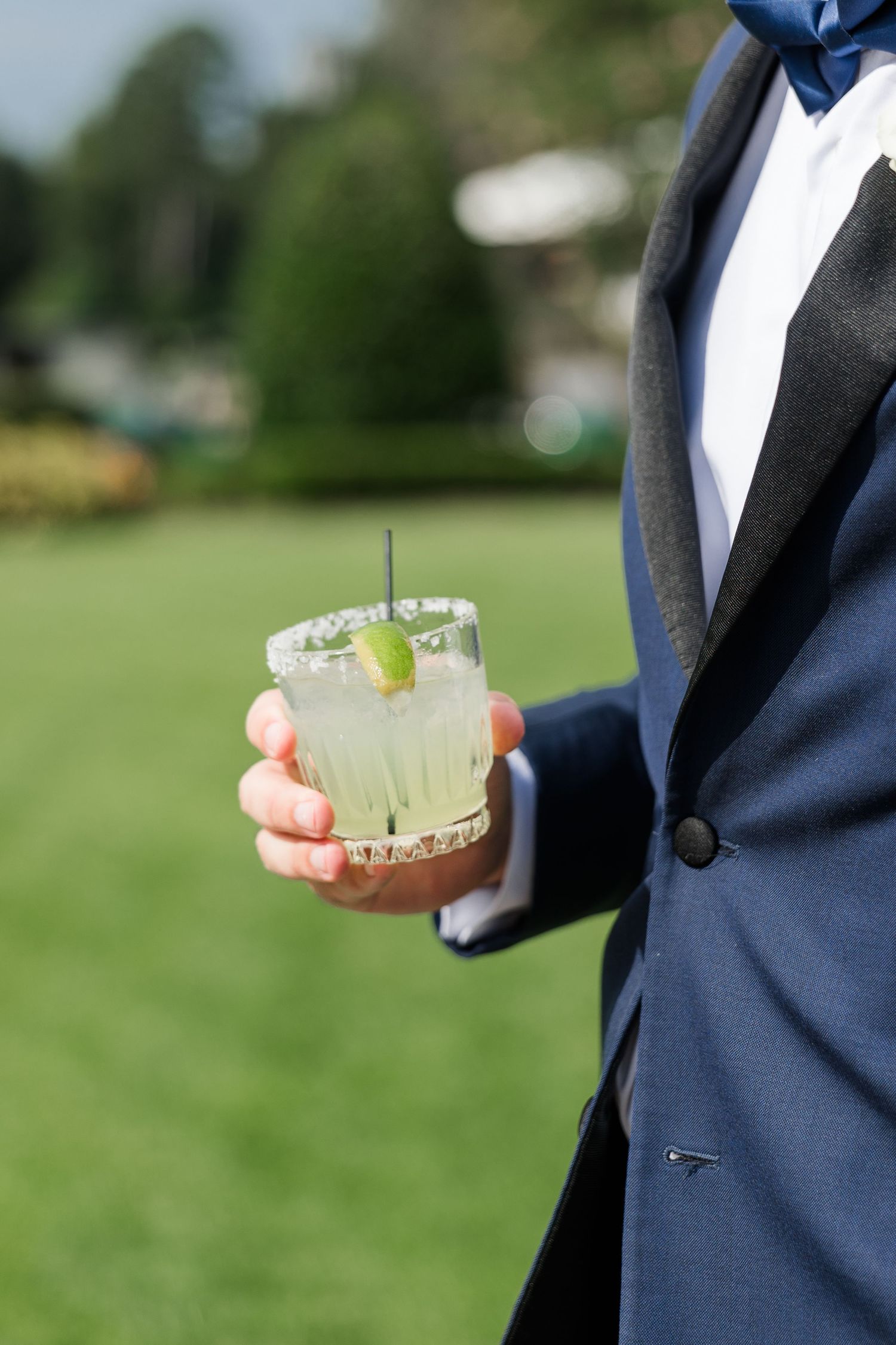 Groom in navy tuxedo holding a margarita cocktail with lime garnish at an outdoor wedding reception.