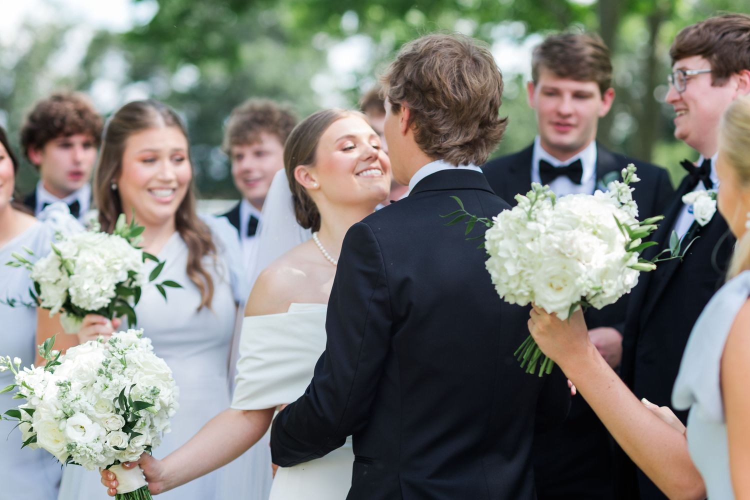Bride and groom share a romantic moment surrounded by cheering wedding party holding white bouquets.
