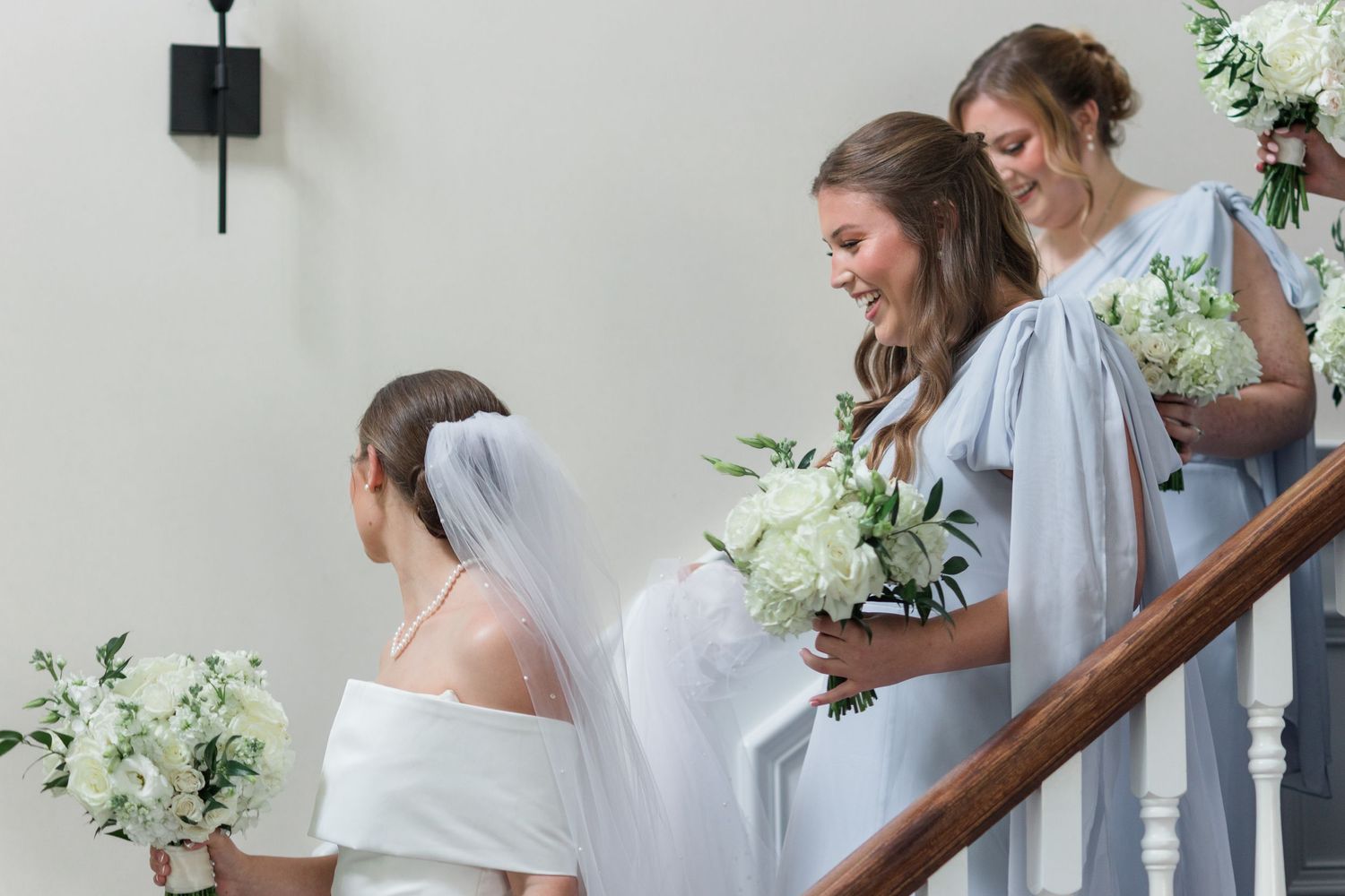 Bride in white gown and veil laughs with bridesmaids in blue dresses on elegant staircase.