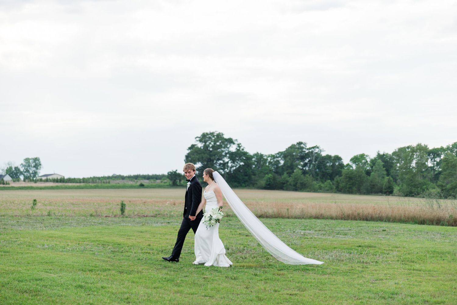 Bride and groom walk hand in hand across a lush green field with a scenic countryside backdrop on their wedding day.