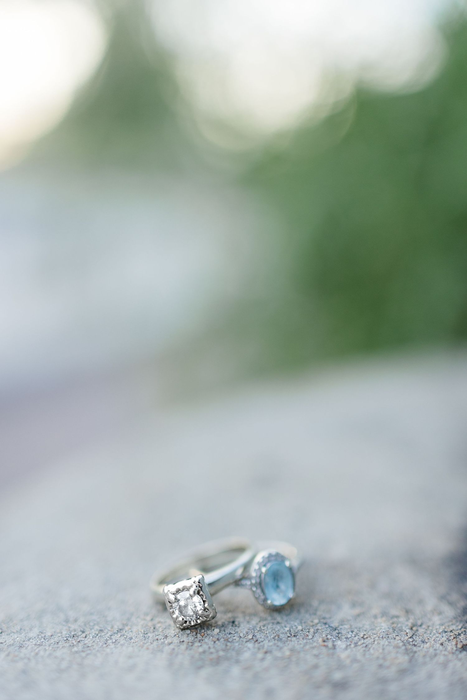 Two elegant silver engagement rings, one with a diamond and one with a blue stone, rest on a stone surface.