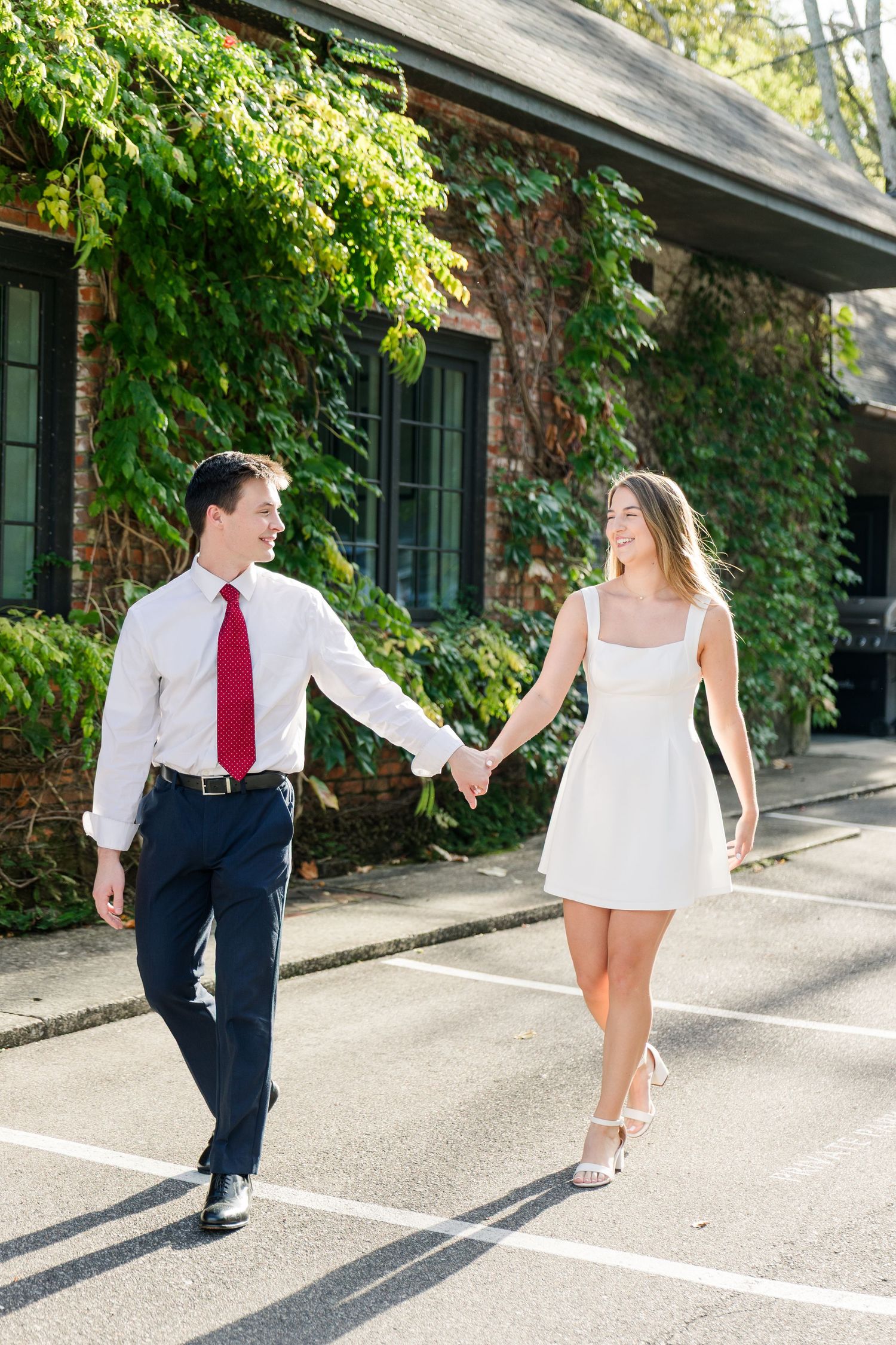 Happy couple holding hands and walking together outside ivy-covered brick building on sunny day.
