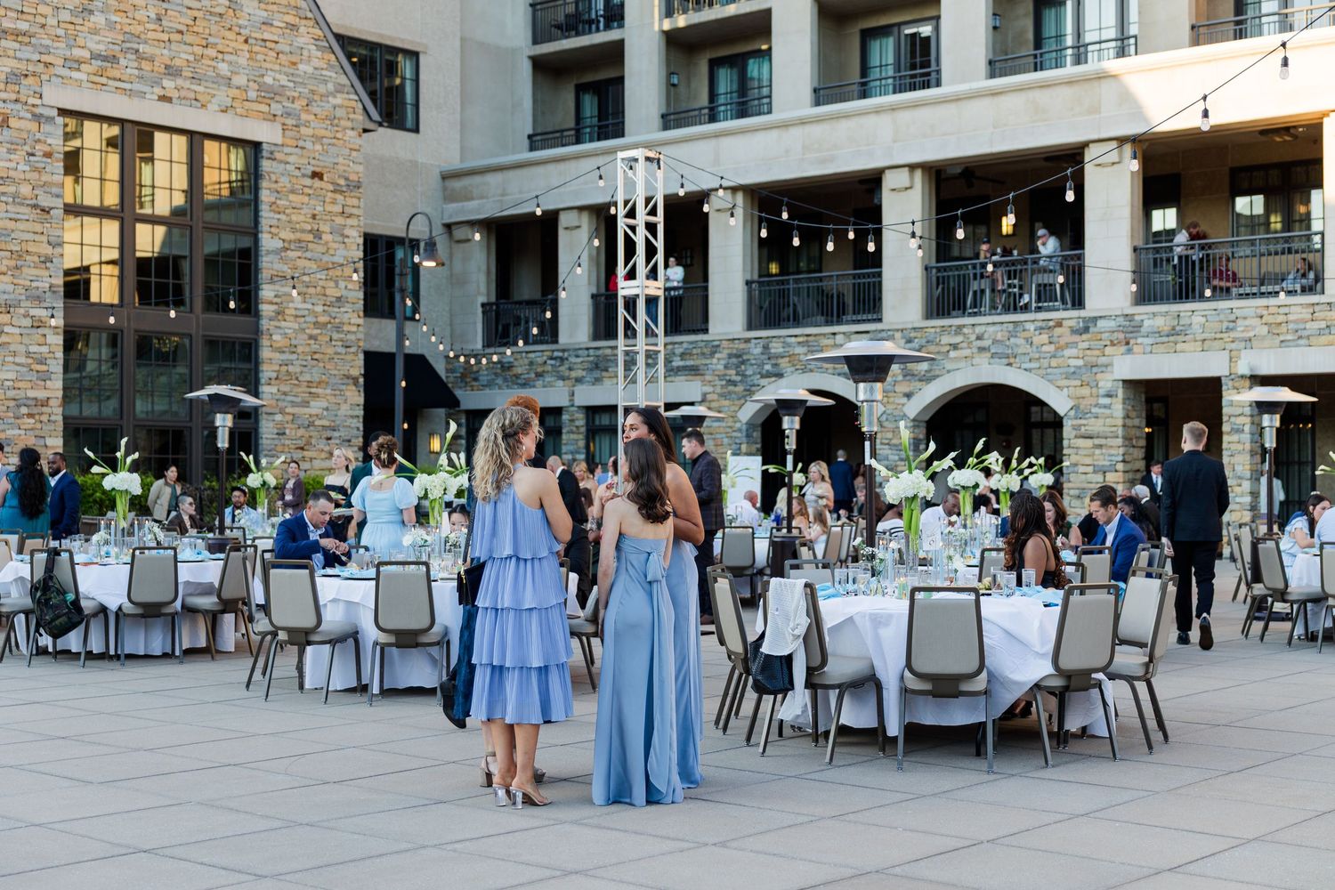 Elegant outdoor wedding reception with round tables, floral centerpieces, and guests in blue dresses at a stone venue.