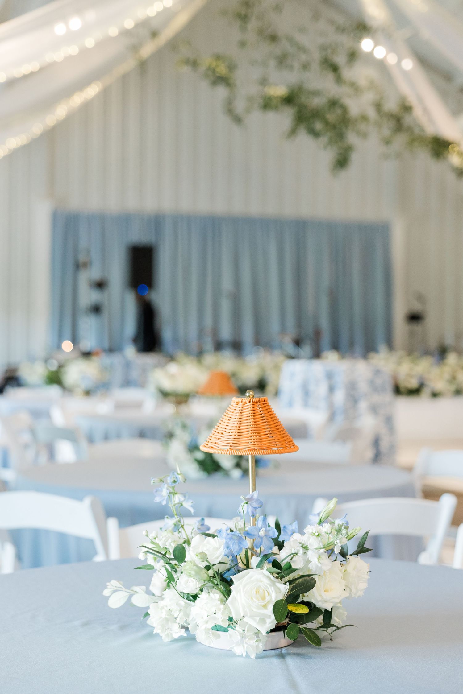 Elegant wedding reception with white floral centerpiece, small wicker lamp, blue tablecloths, and white chairs in a decorated venue.