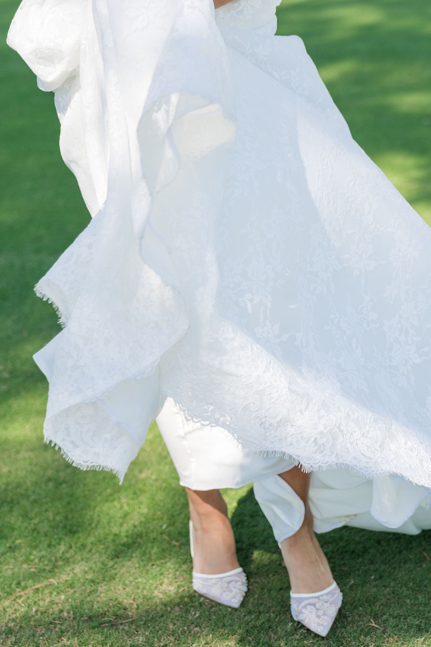 Bride in a flowing white lace wedding dress and white heels standing on green grass outdoors.