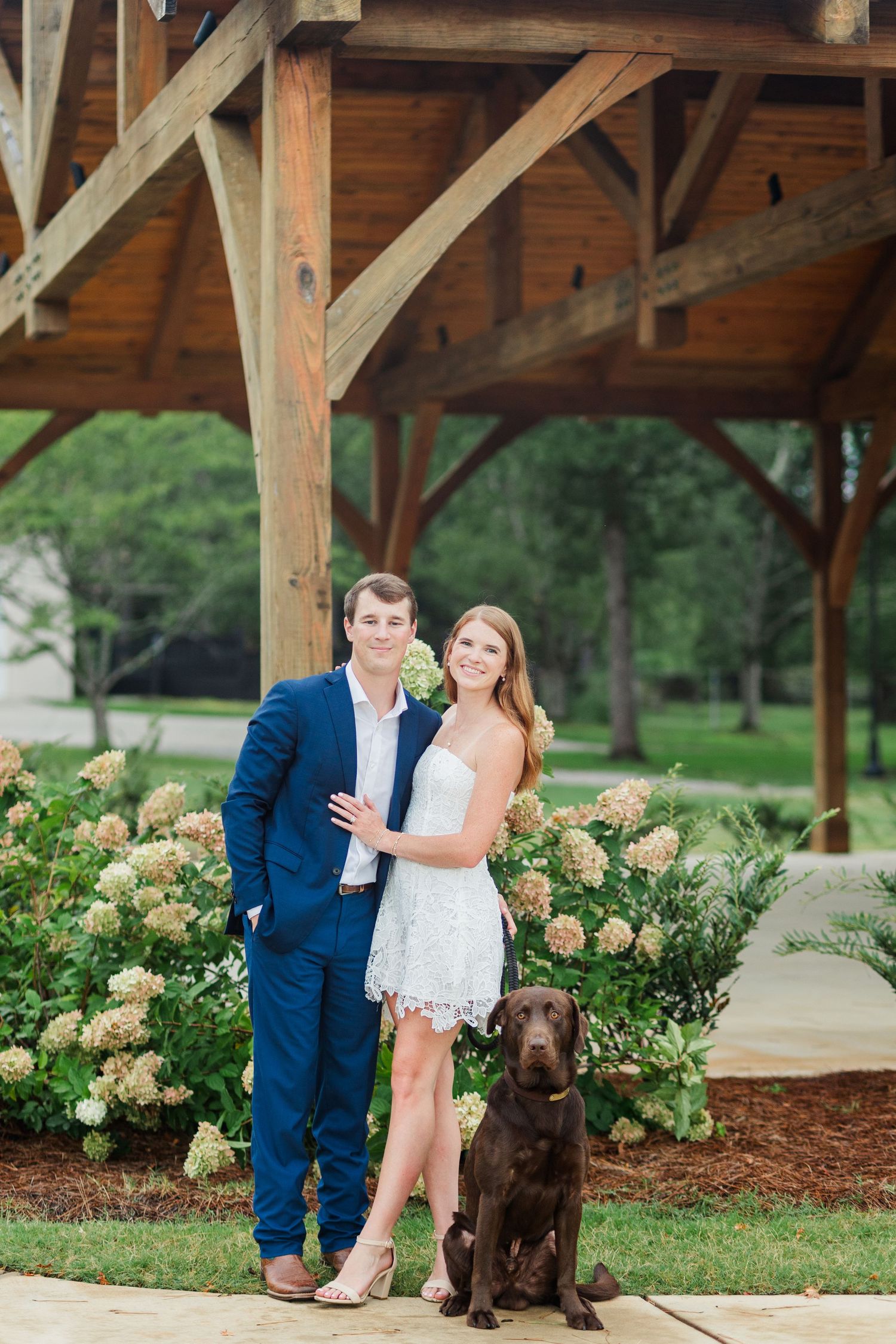 Couple in blue suit and white lace dress posing with chocolate Labrador under rustic wooden pavilion.