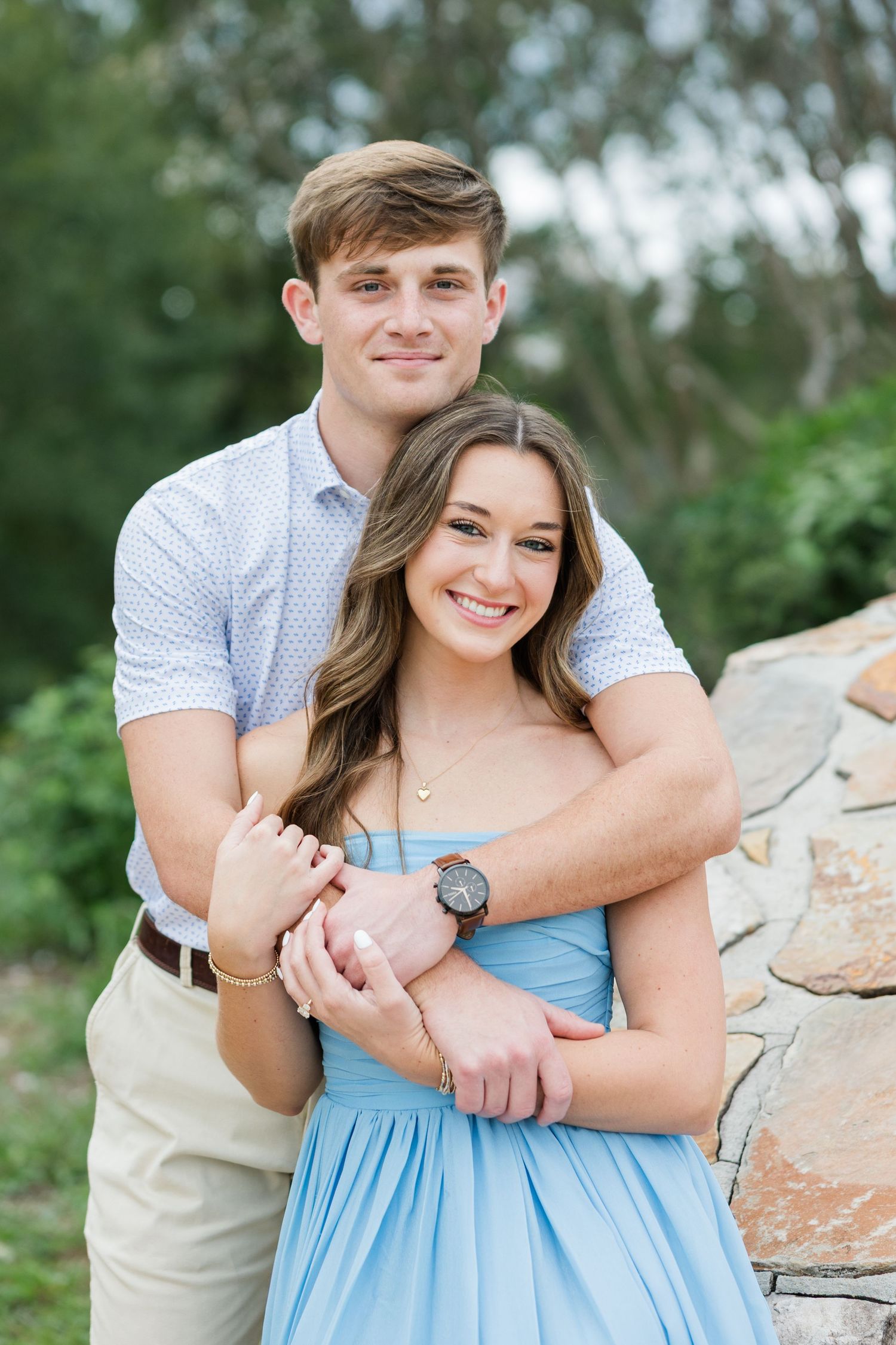 A smiling young couple embracing outdoors, the man hugging the woman in a blue dress from behind beside a stone wall.