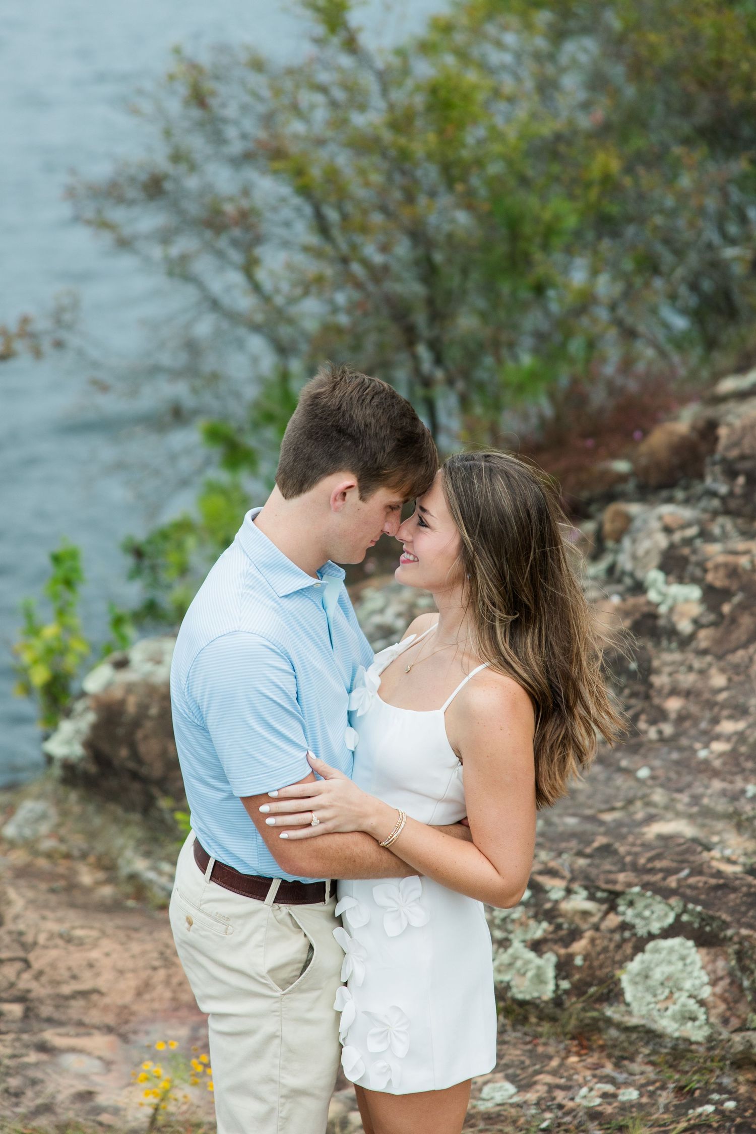 Couple embracing forehead to forehead on a rocky lakeside overlook, she in a white dress and he in a light blue polo.