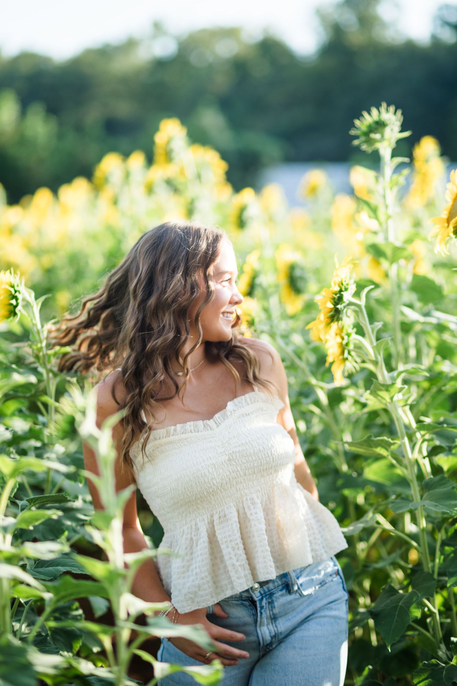 A smiling young woman in a white crop top and jeans stands among tall sunflowers in a sunny field.