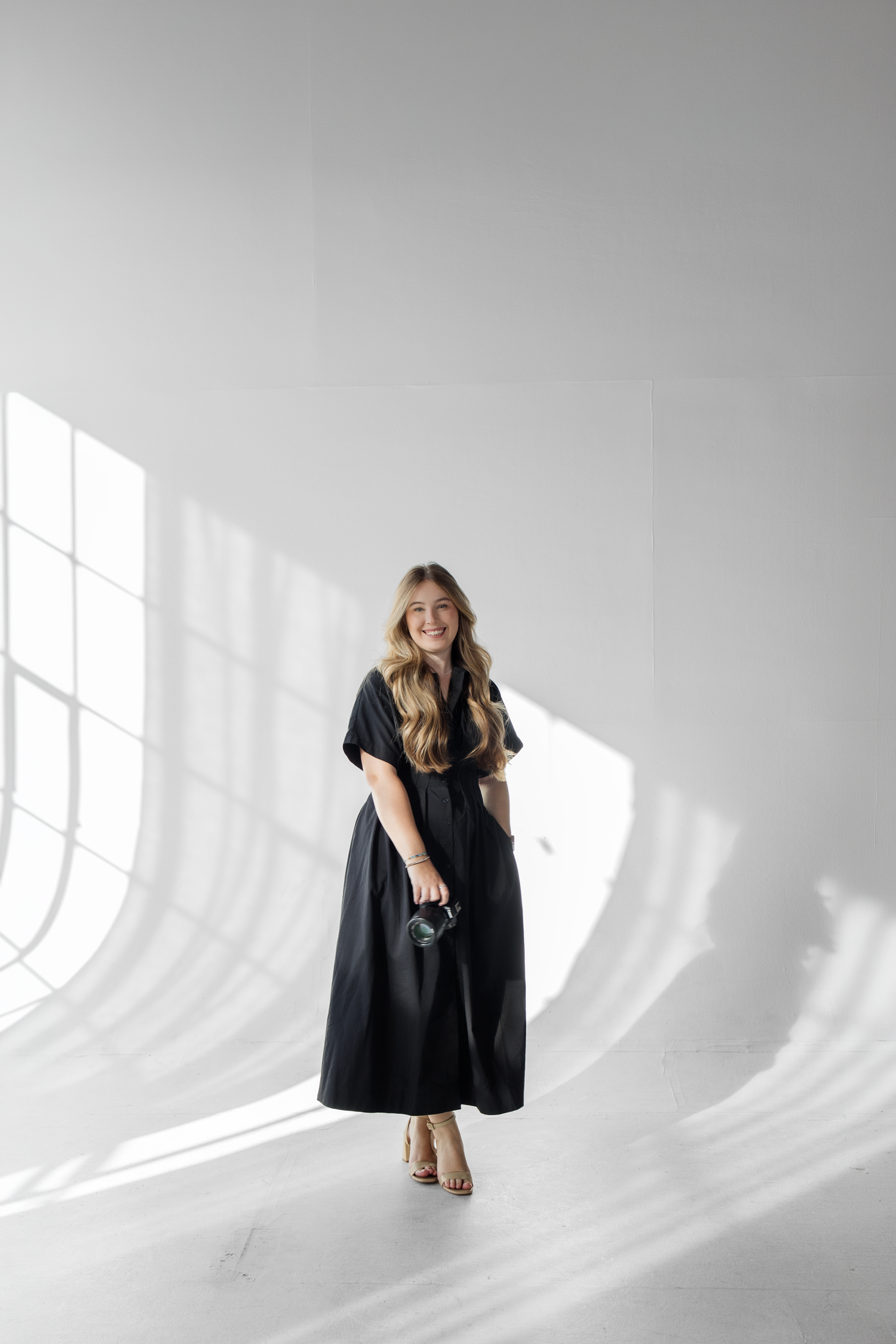 Woman in black maxi dress and heels smiling in bright white studio with natural light shadows.