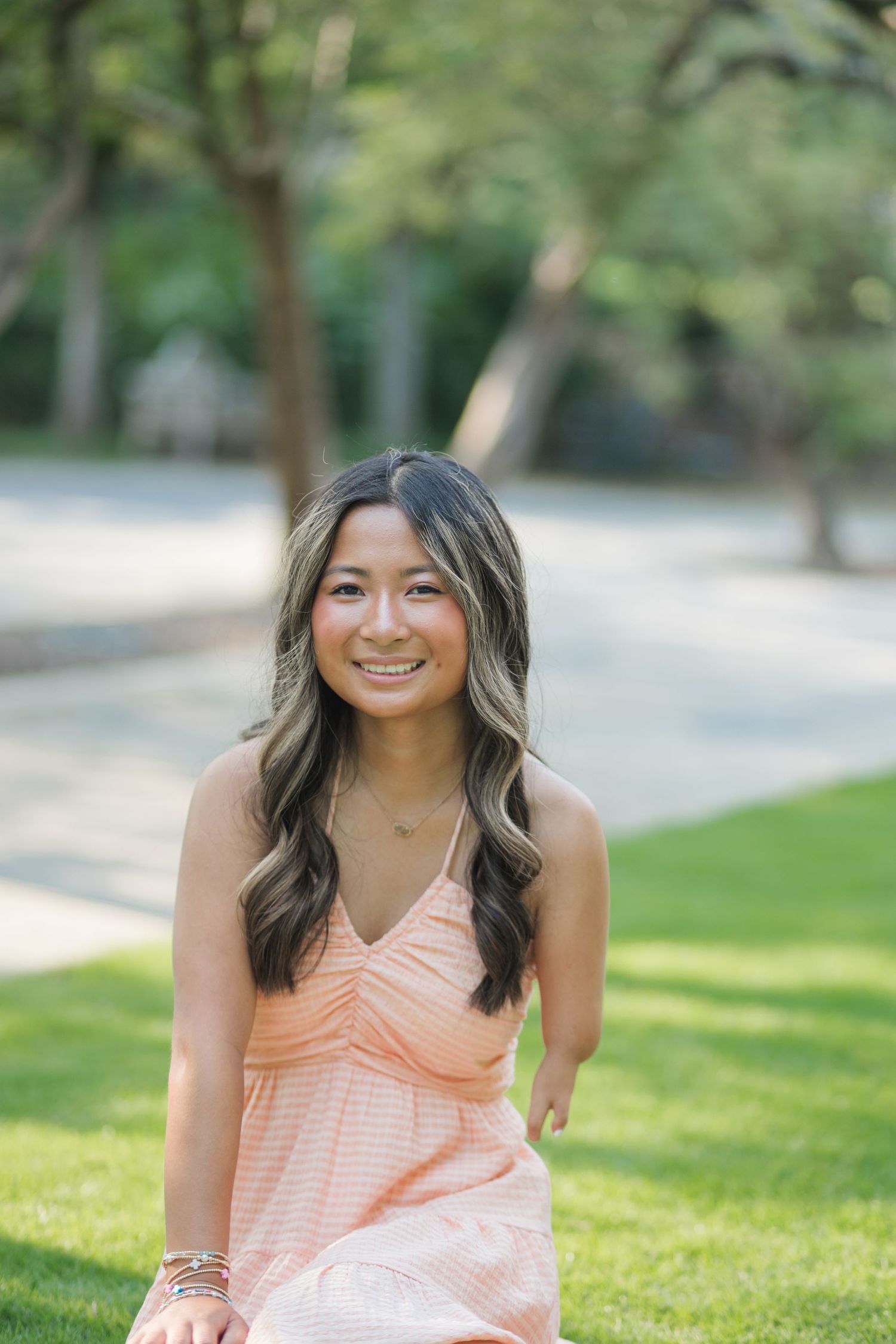 Young woman in peach sundress smiling outdoors on green grass with trees in background.
