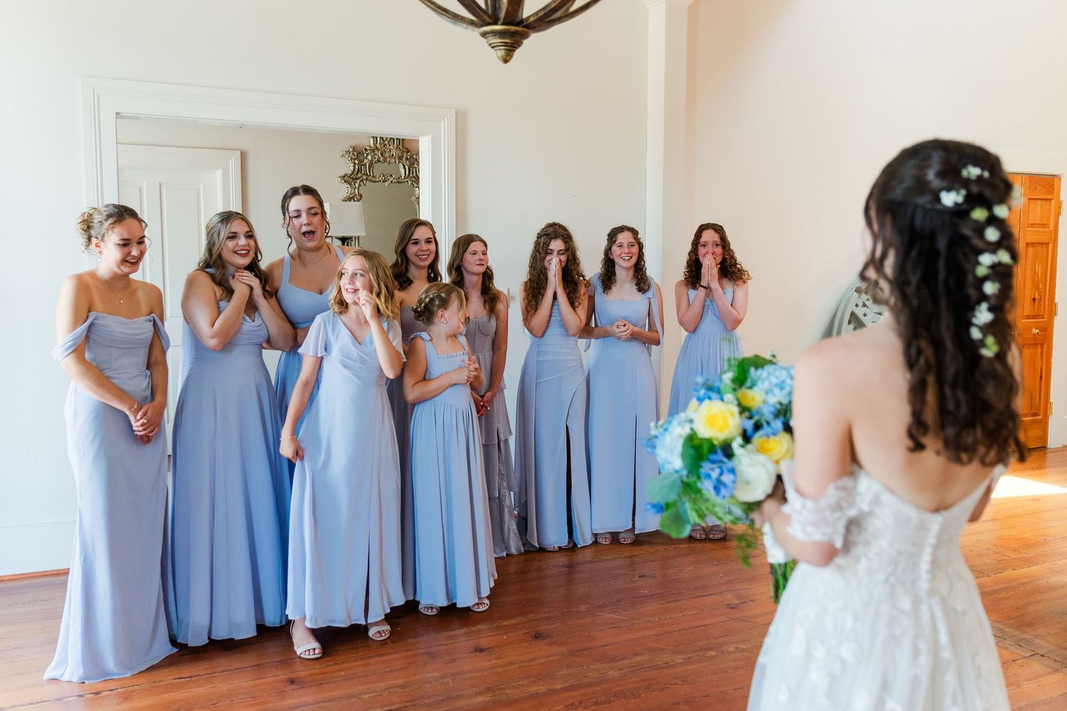 Bridesmaids in blue dresses react with joy and tears seeing bride for first time during wedding first look.