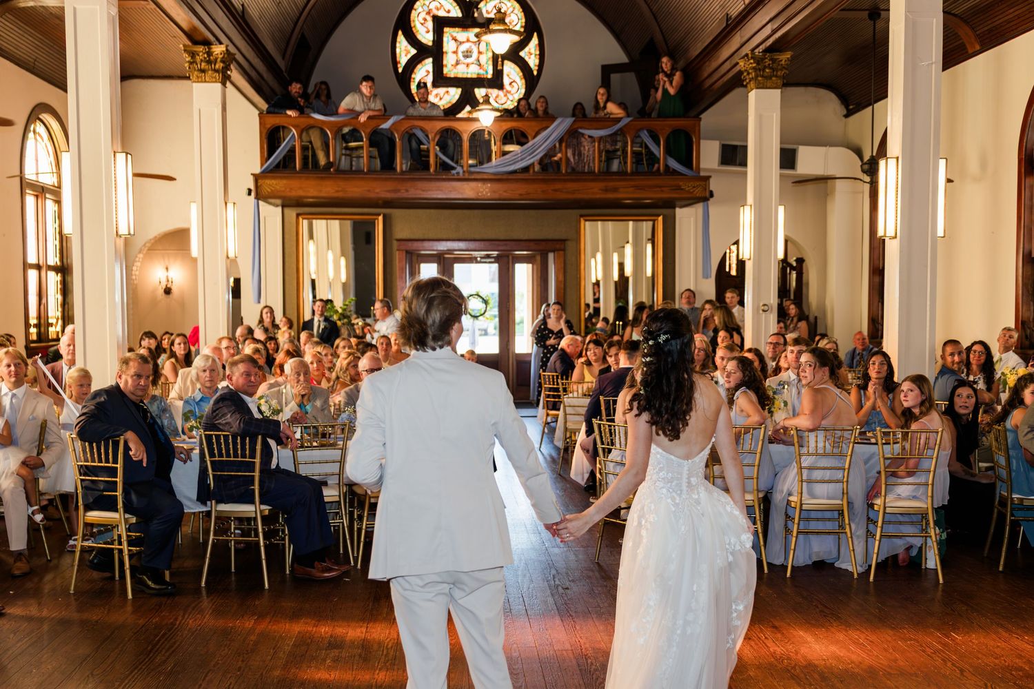Bride and groom entering elegant wedding reception hall with guests seated at decorated tables.
