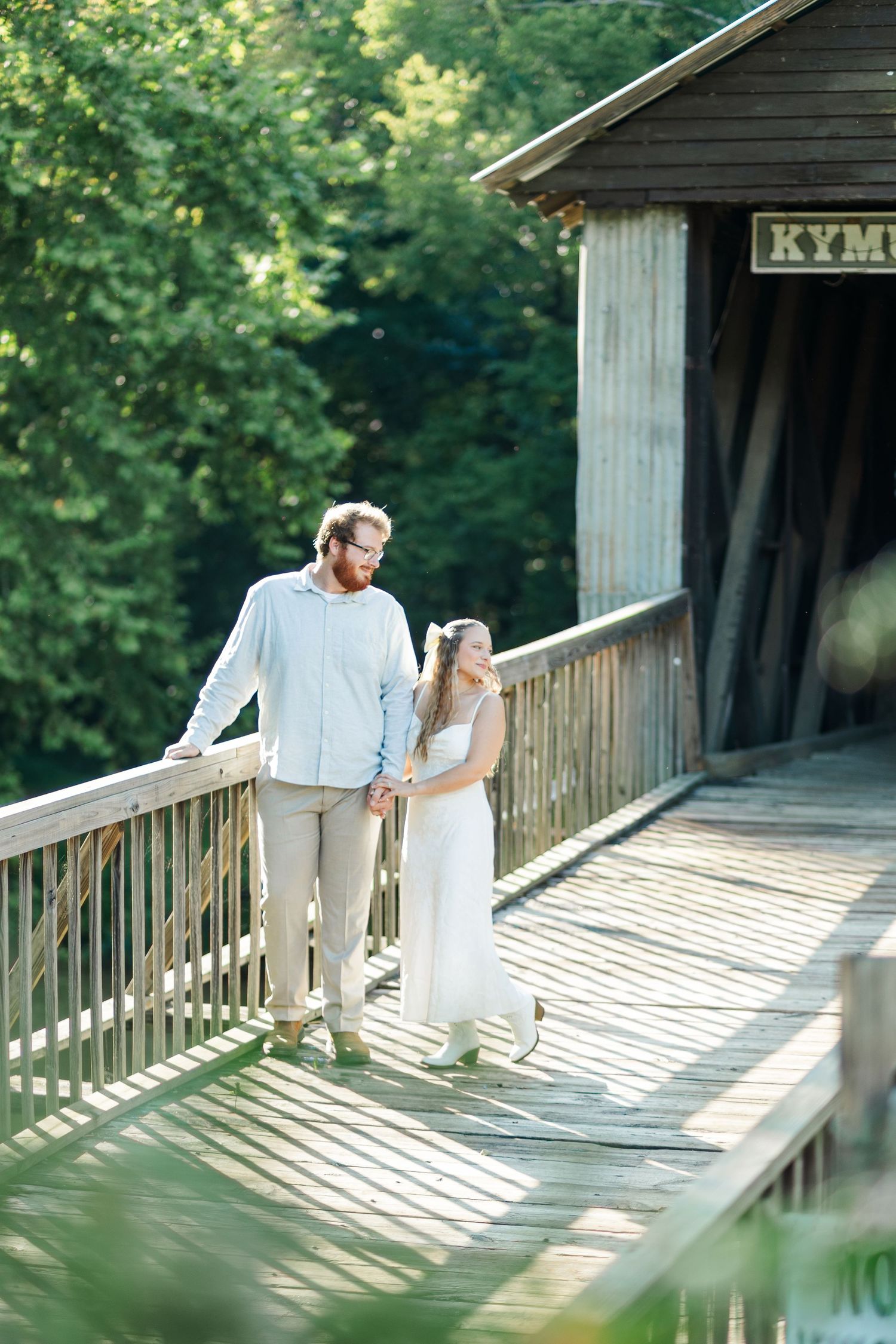 Couple holding hands walking on wooden covered bridge surrounded by lush green trees in summer.