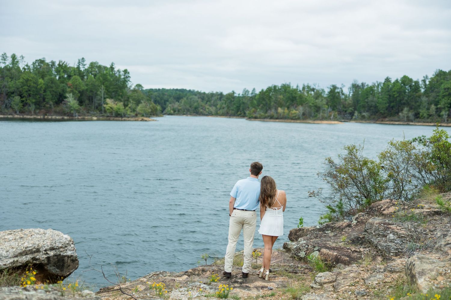 Couple standing on a rocky shoreline looking out over a calm lake surrounded by green trees under a cloudy sky.