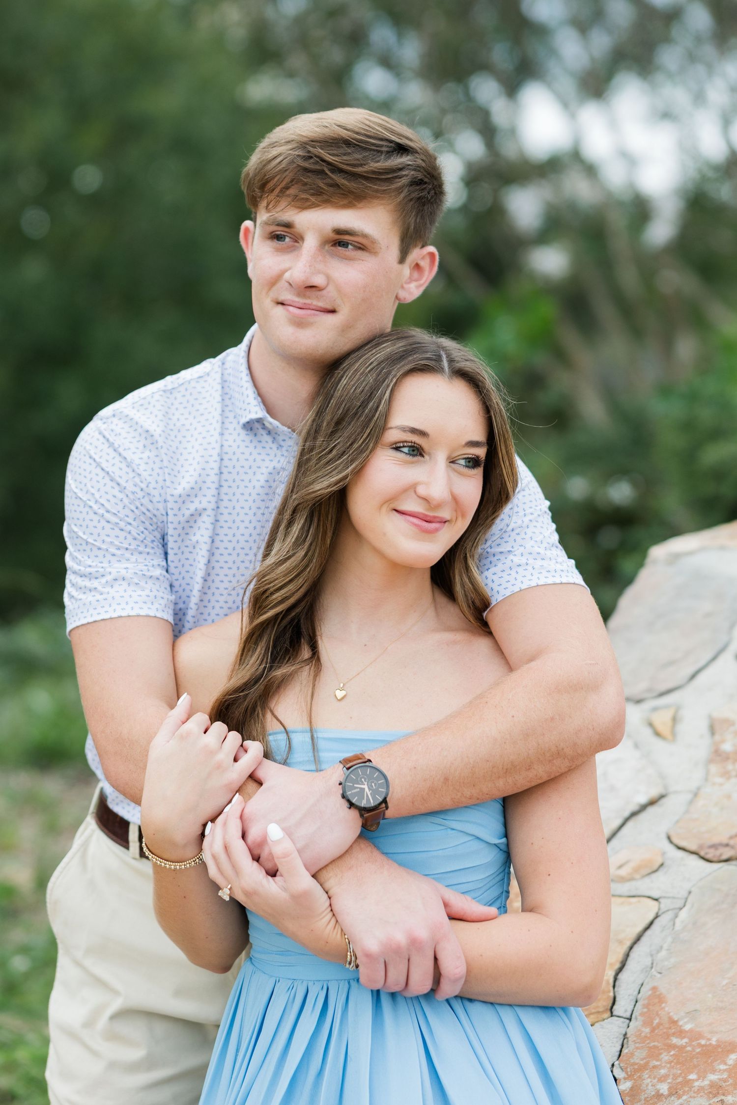 Smiling couple embracing outdoors as a young man in a patterned shirt hugs a woman in a blue dress from behind near a stone wall.