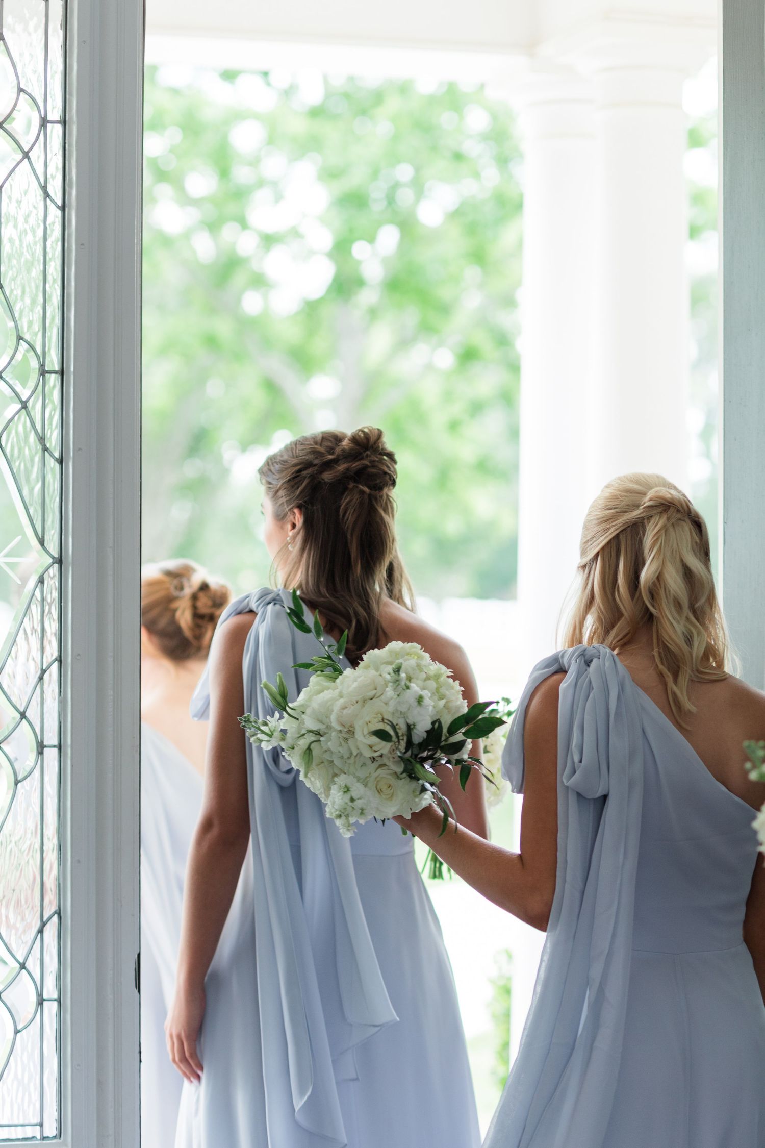 Bridesmaids in dusty blue one-shoulder gowns holding white floral bouquets walk toward elegant venue doors.