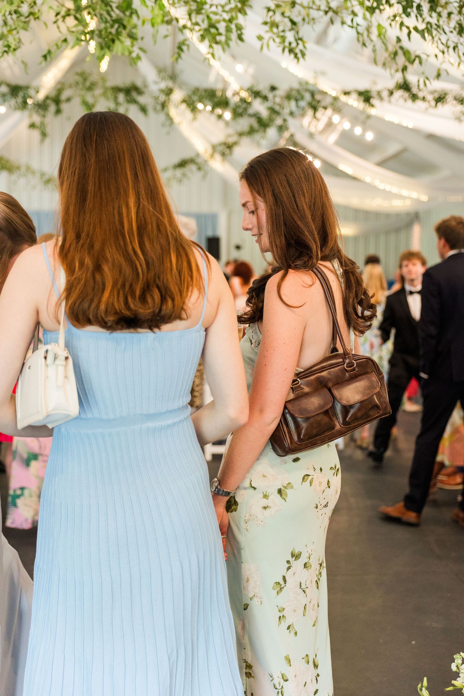 Two young women in blue and floral dresses mingle under decorated tent canopy at wedding reception.