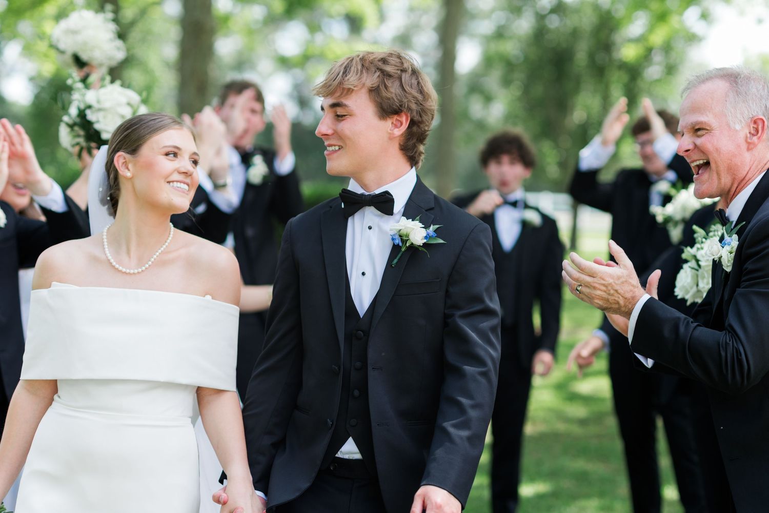 Smiling newlyweds walk hand in hand past cheering wedding party in black tuxedos at outdoor ceremony.