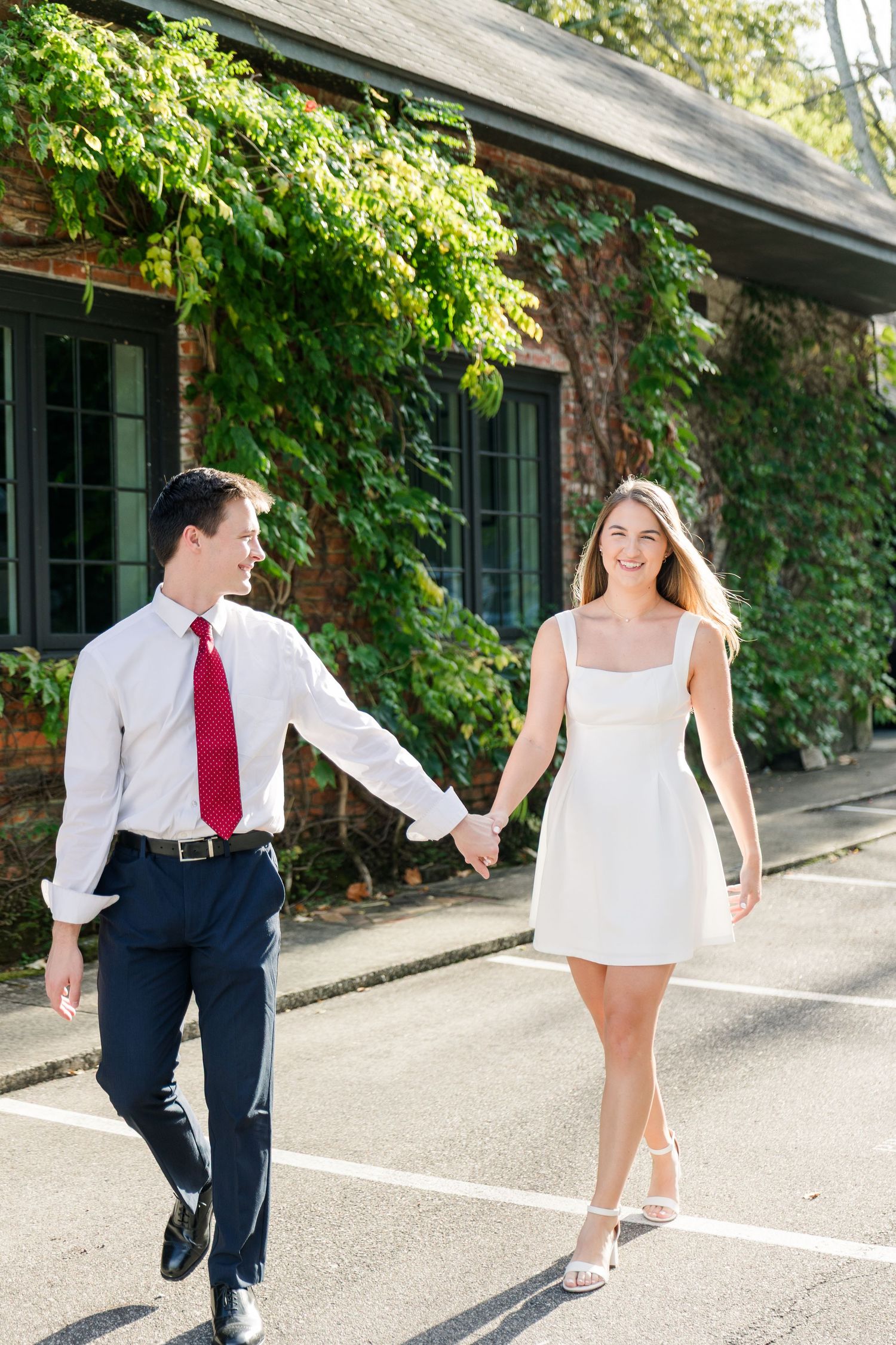 A smiling couple holds hands walking outdoors, man in white shirt and red tie, woman in white mini dress.