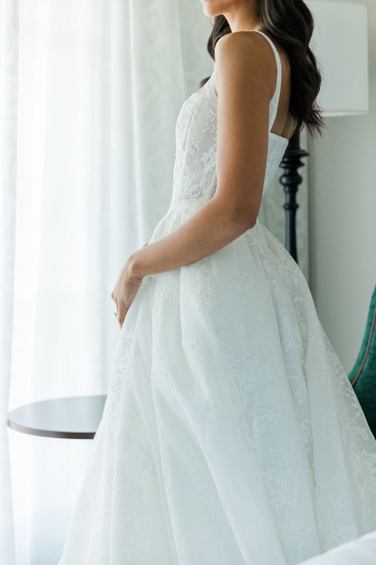 Bride in elegant white lace ball gown wedding dress standing by sheer curtains, back view showing intricate floral details.