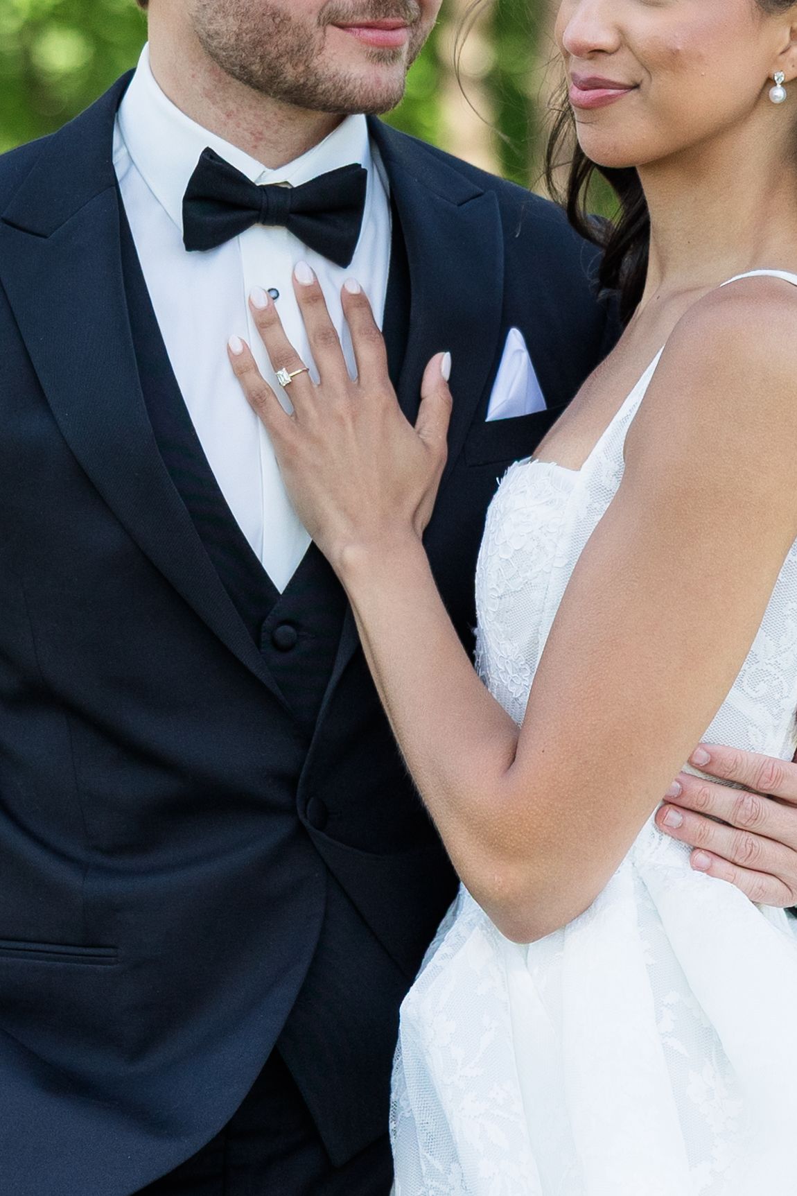 Bride in white lace gown rests hand on groom's tuxedo chest, showcasing diamond engagement ring at wedding.