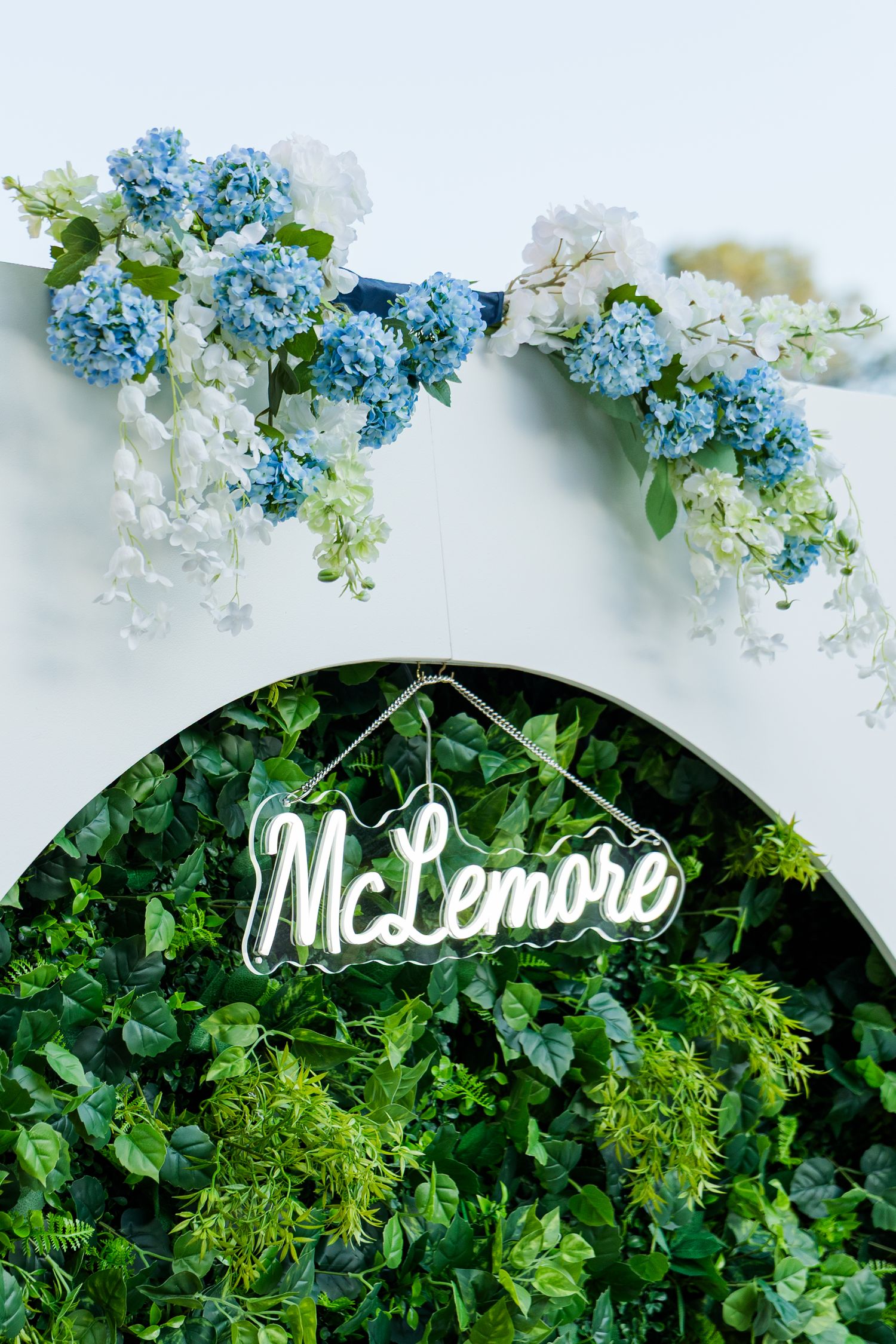 McLemore neon sign hanging on a lush green circular hedge wall with blue and white hydrangea floral arrangement above.