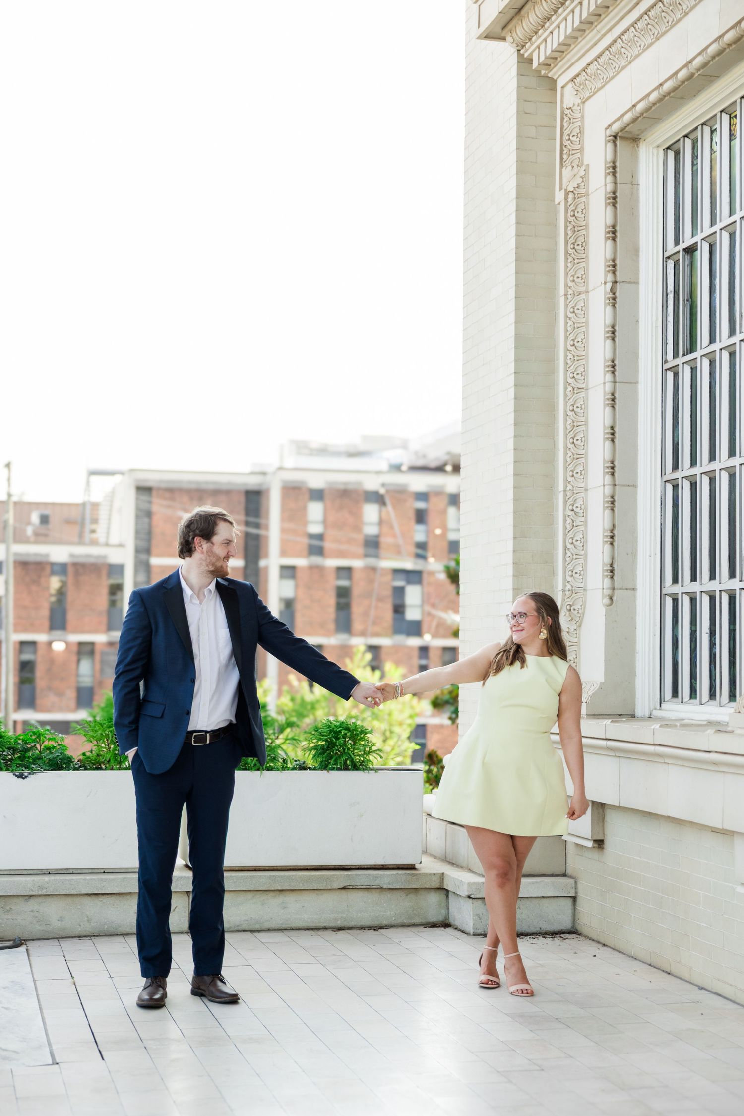 Couple holding hands on a marble terrace, man in navy suit and woman in yellow dress, smiling at each other outdoors.