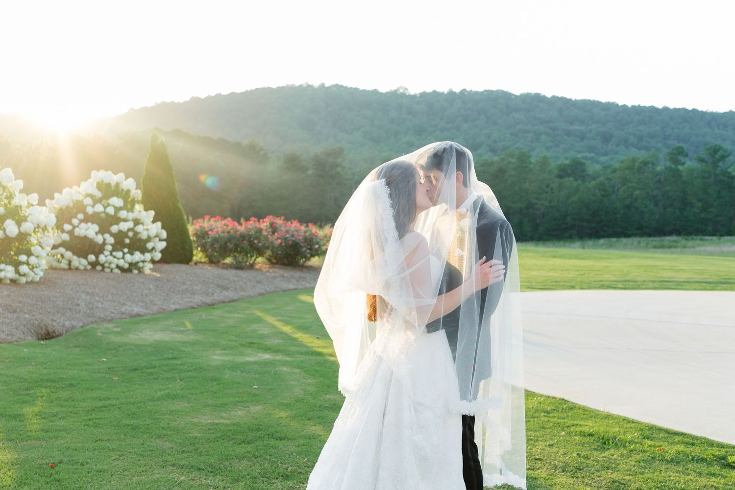 Bride and groom share a kiss under a veil at sunset with lush green hills and white flowers in the background.