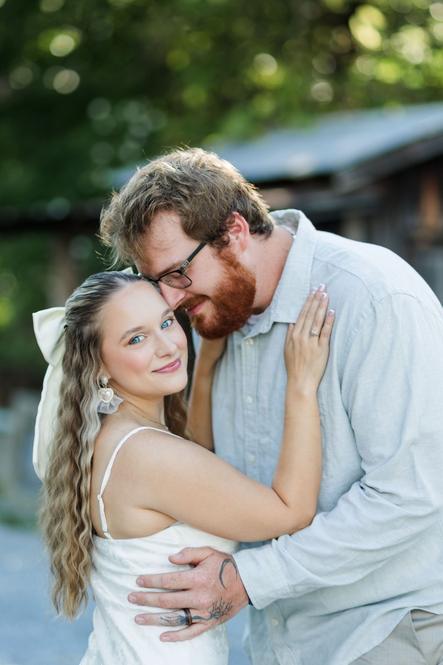 Couple embracing outdoors, woman in white dress with bow smiling at camera, bearded man with glasses nuzzling her head.