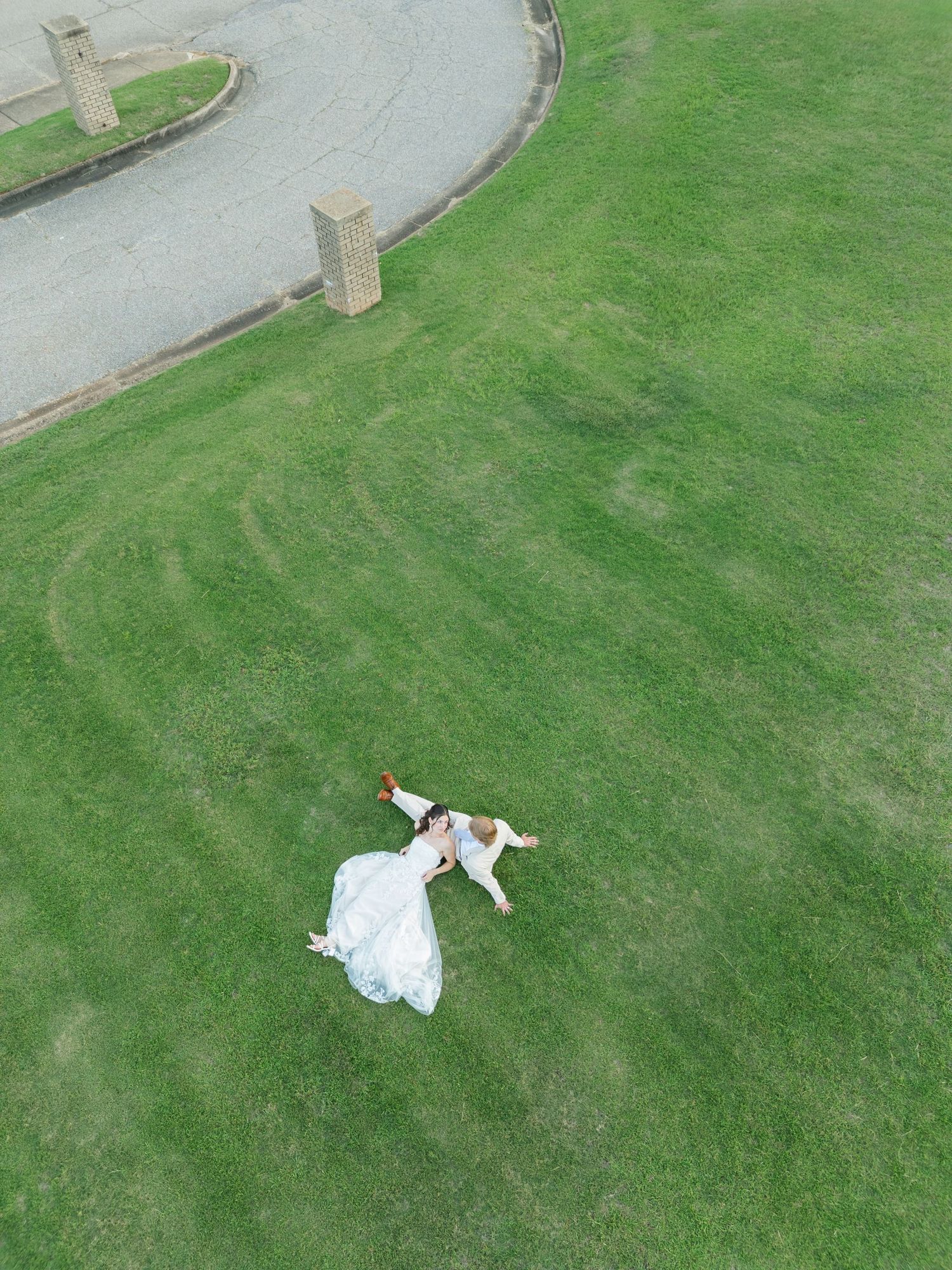 Aerial drone shot of bride and groom lying together on a lush green lawn in their wedding attire.
