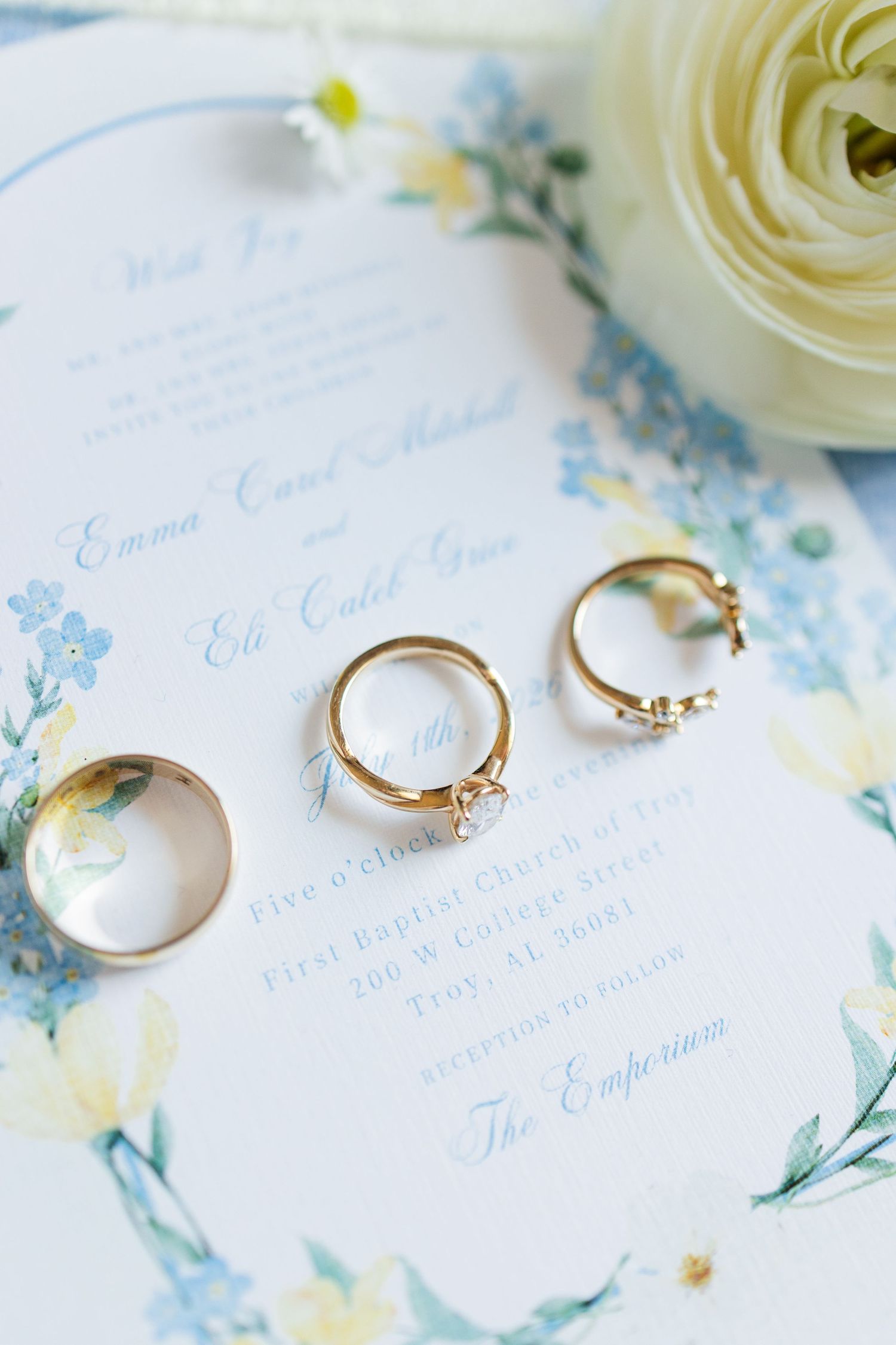 Gold wedding rings and engagement ring displayed on floral blue and white wedding invitation with white rose.