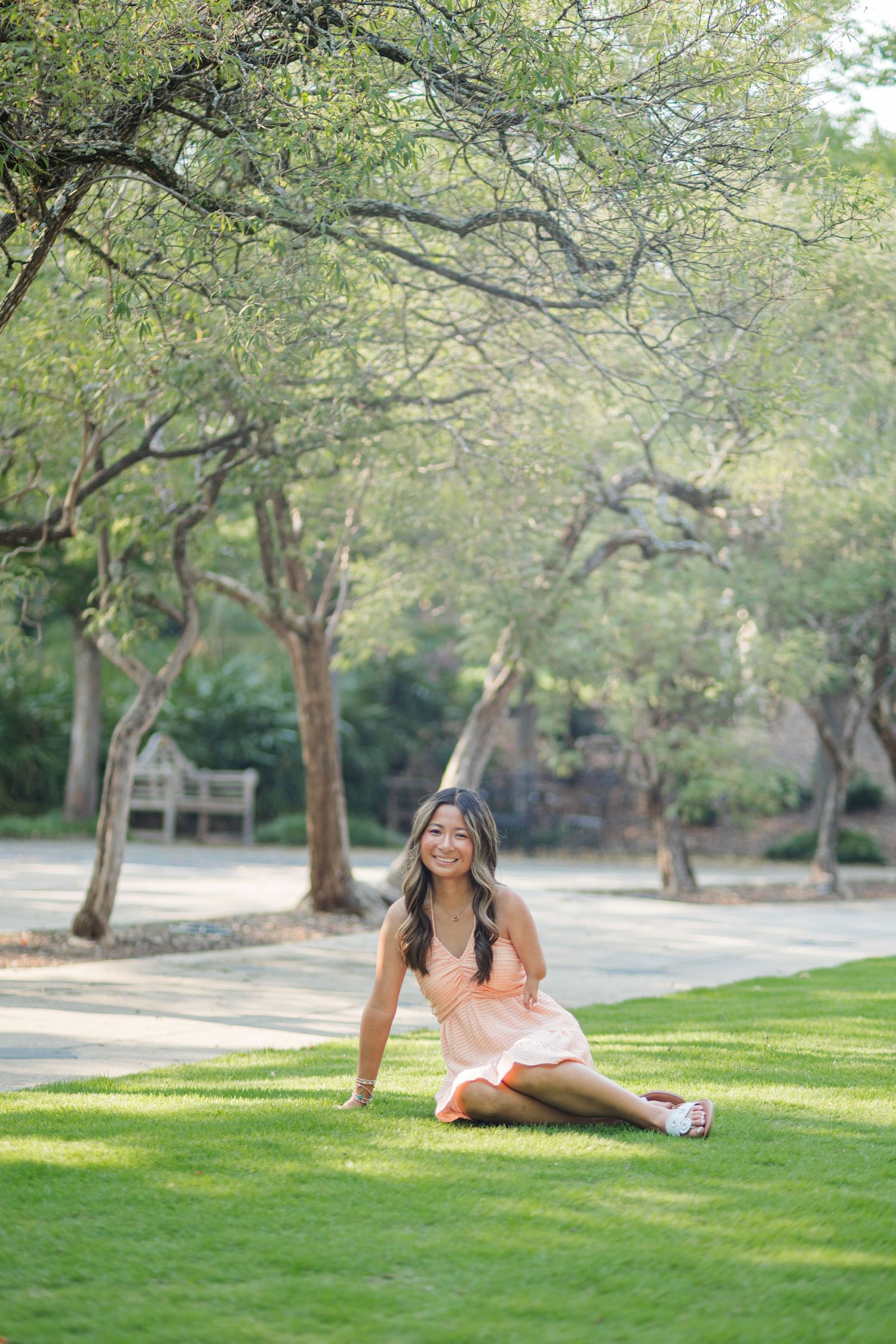 Young woman in peach dress sitting on grass beneath green trees in sunny park.