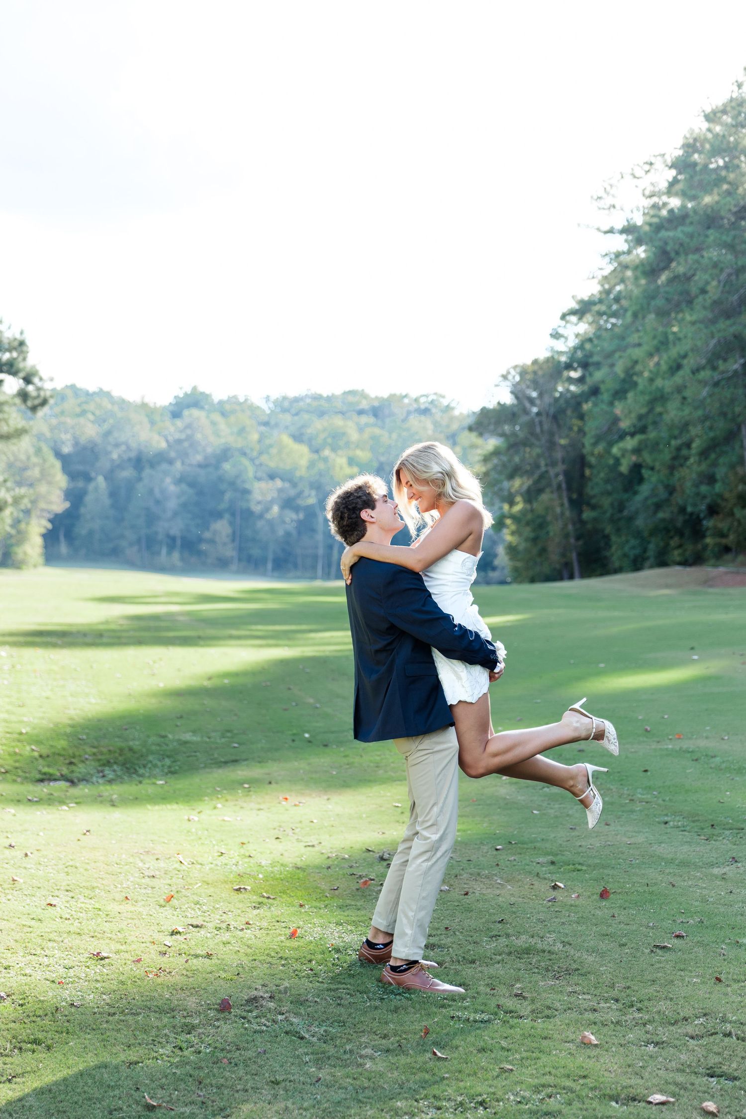 Groom lifting bride on a golf course during romantic engagement photo session outdoors in natural sunlight.