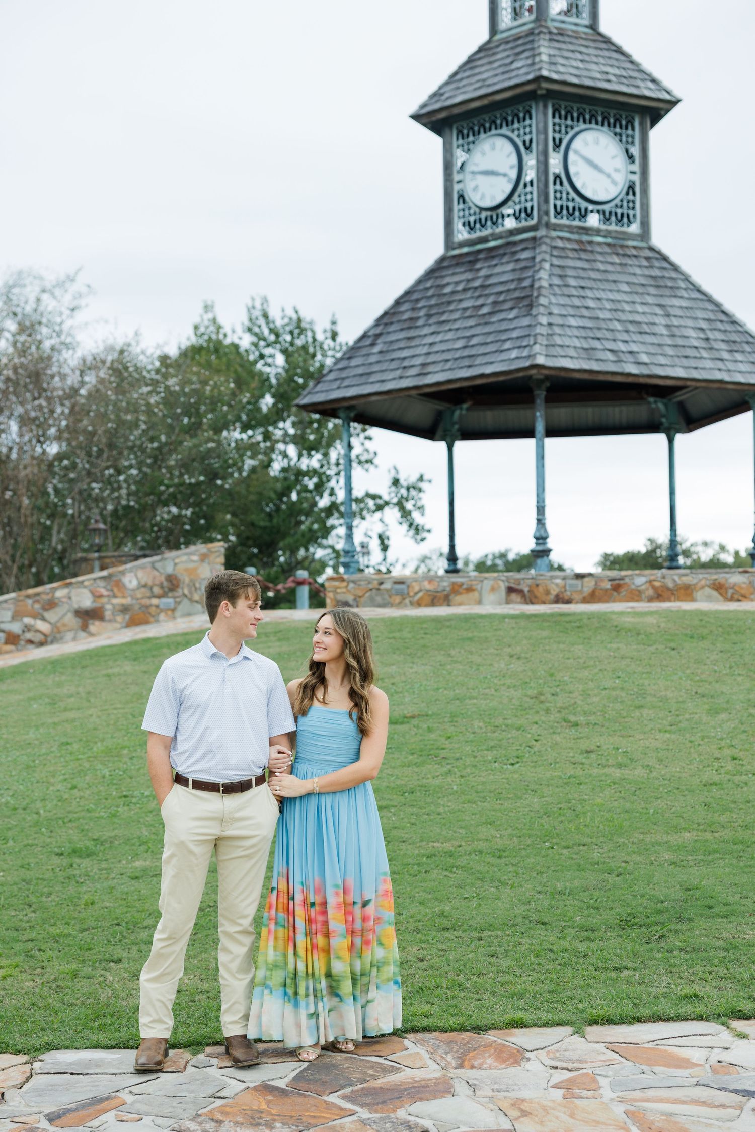 Smiling couple in coordinated outfits embrace on a stone path before a grassy lawn and a pavilion topped with a clock tower.