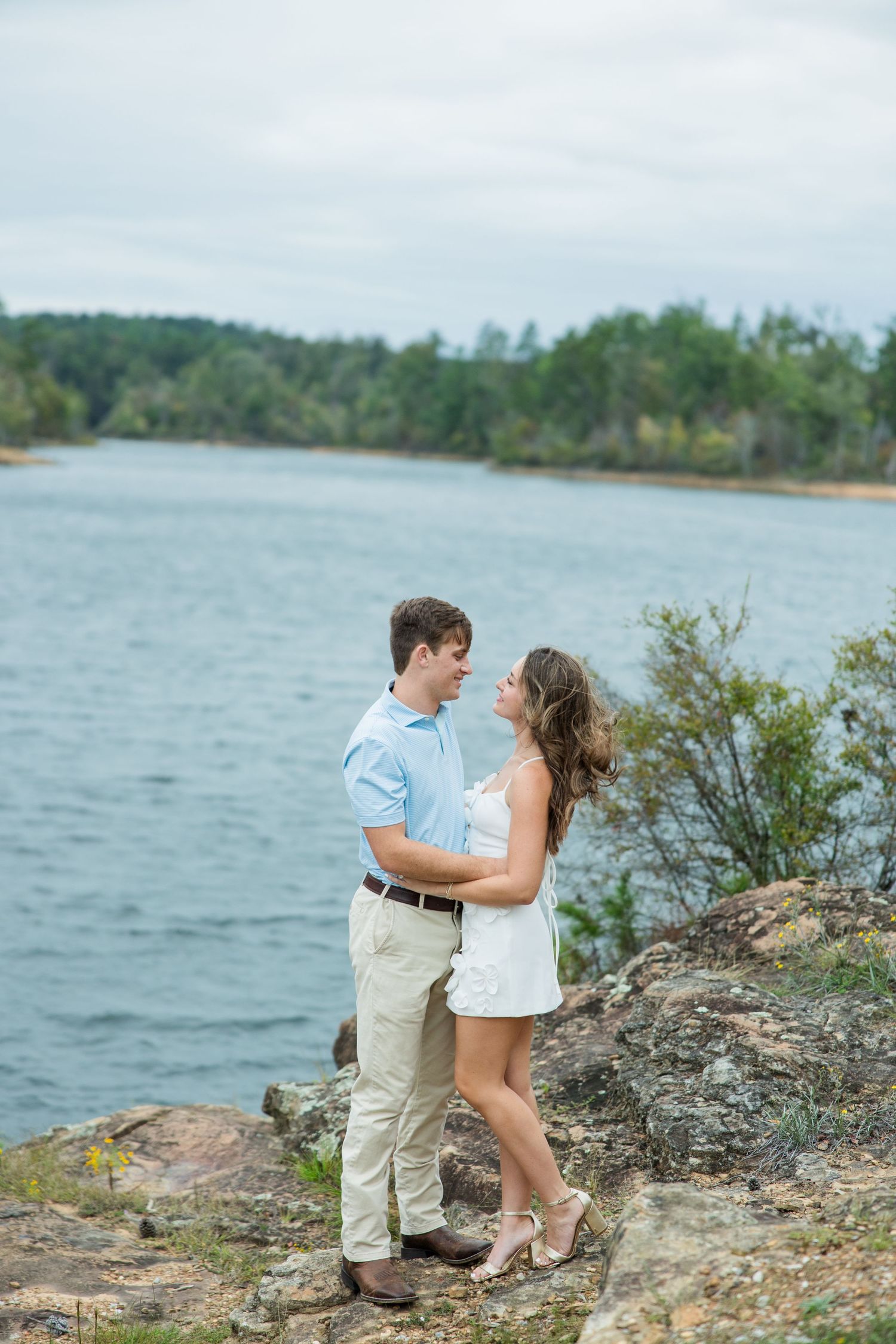 Engaged couple embraces and smiles on a rocky lakeside overlook with calm water and green trees behind them.