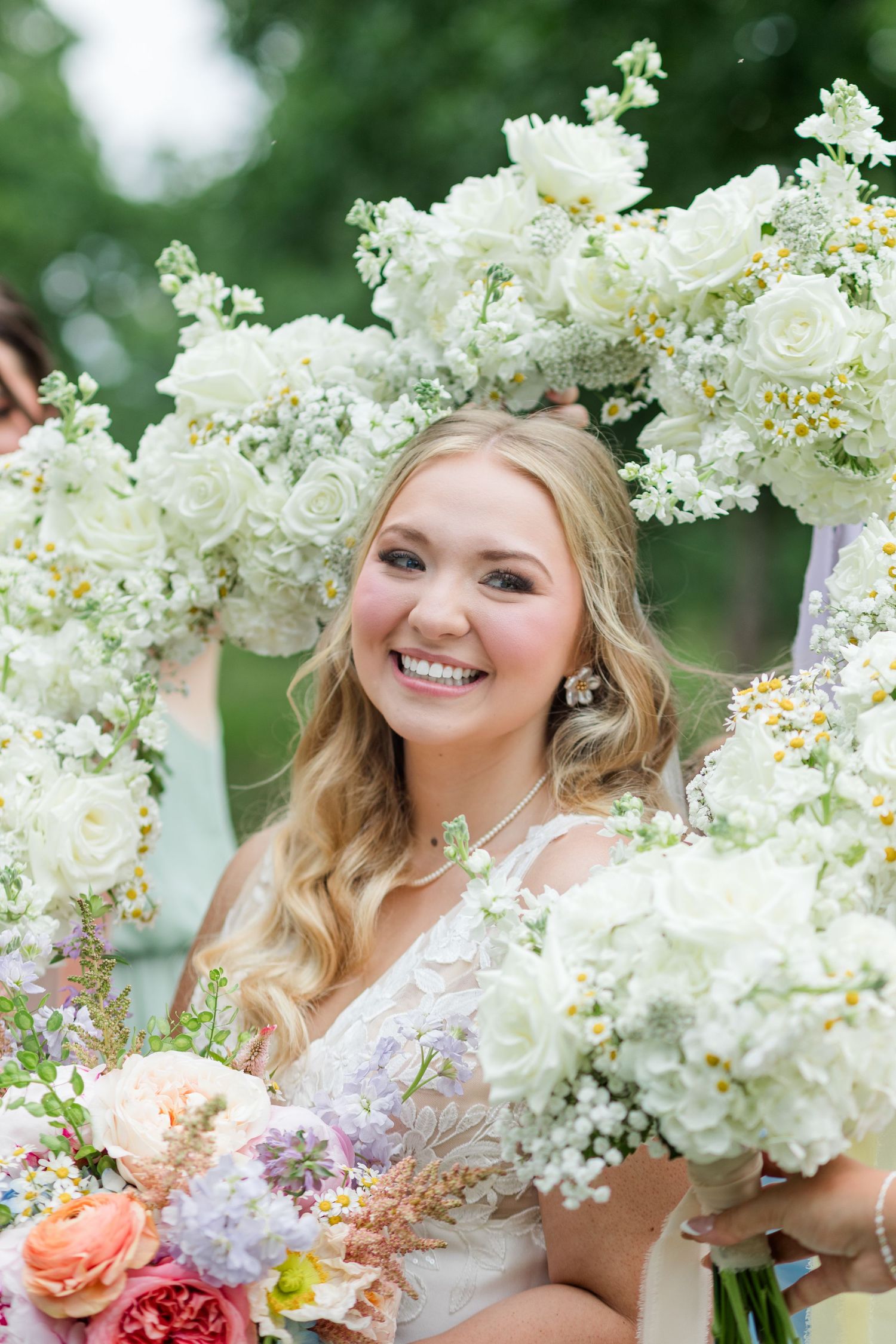 Smiling bride framed by white floral arch holding colorful bouquet at outdoor wedding ceremony.