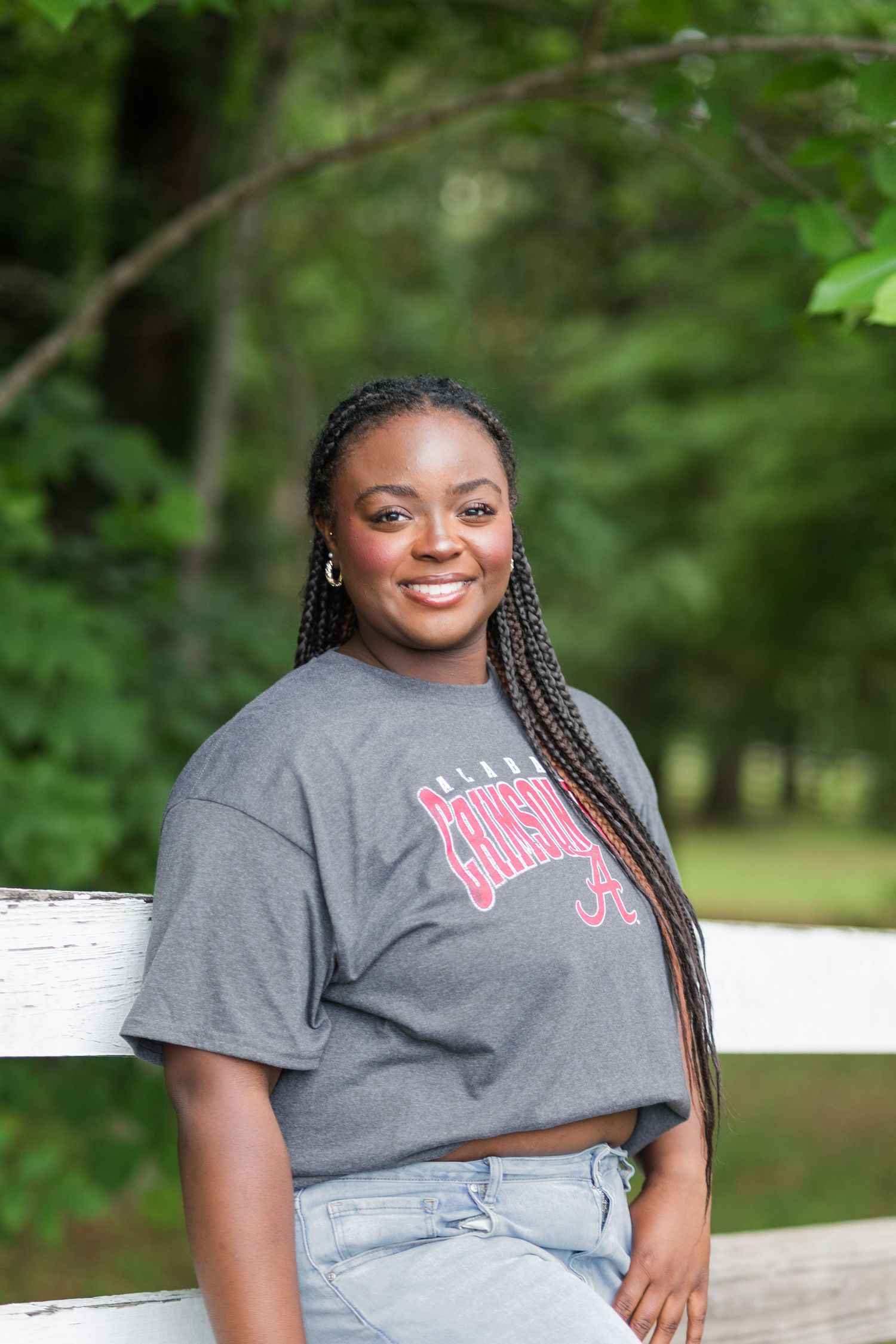 Young woman smiling in Alabama Crimson Tide t-shirt leaning on white fence outdoors.