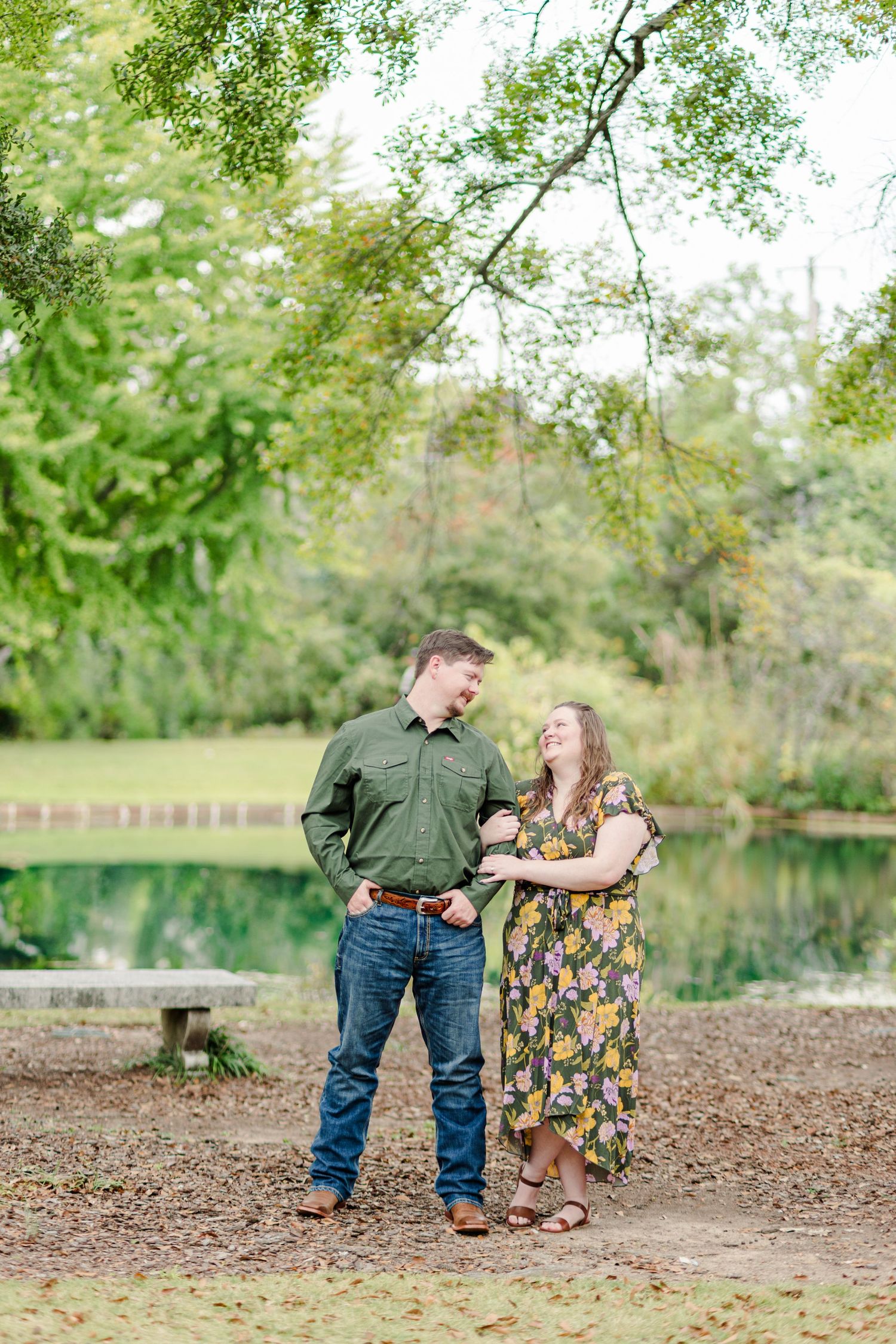 A couple smiles at each other near a pond surrounded by lush green trees during an outdoor engagement session.