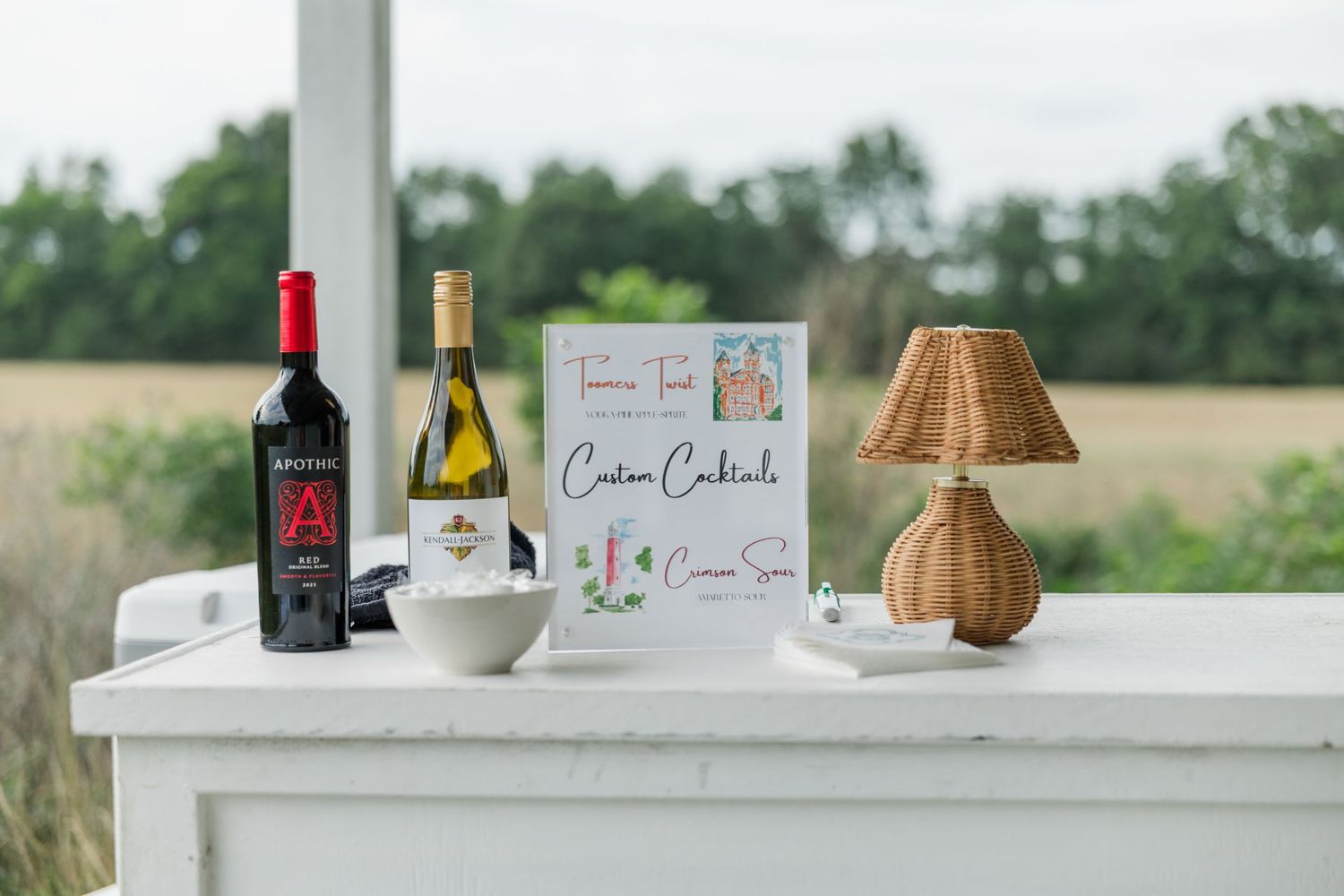 Custom cocktails sign displayed with red and white wine bottles on white bar table outdoors.