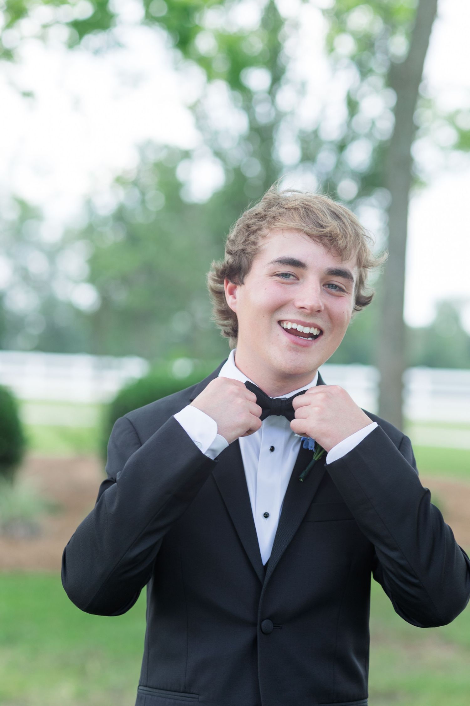 Smiling young man in a black tuxedo adjusting his bow tie outdoors before a wedding.